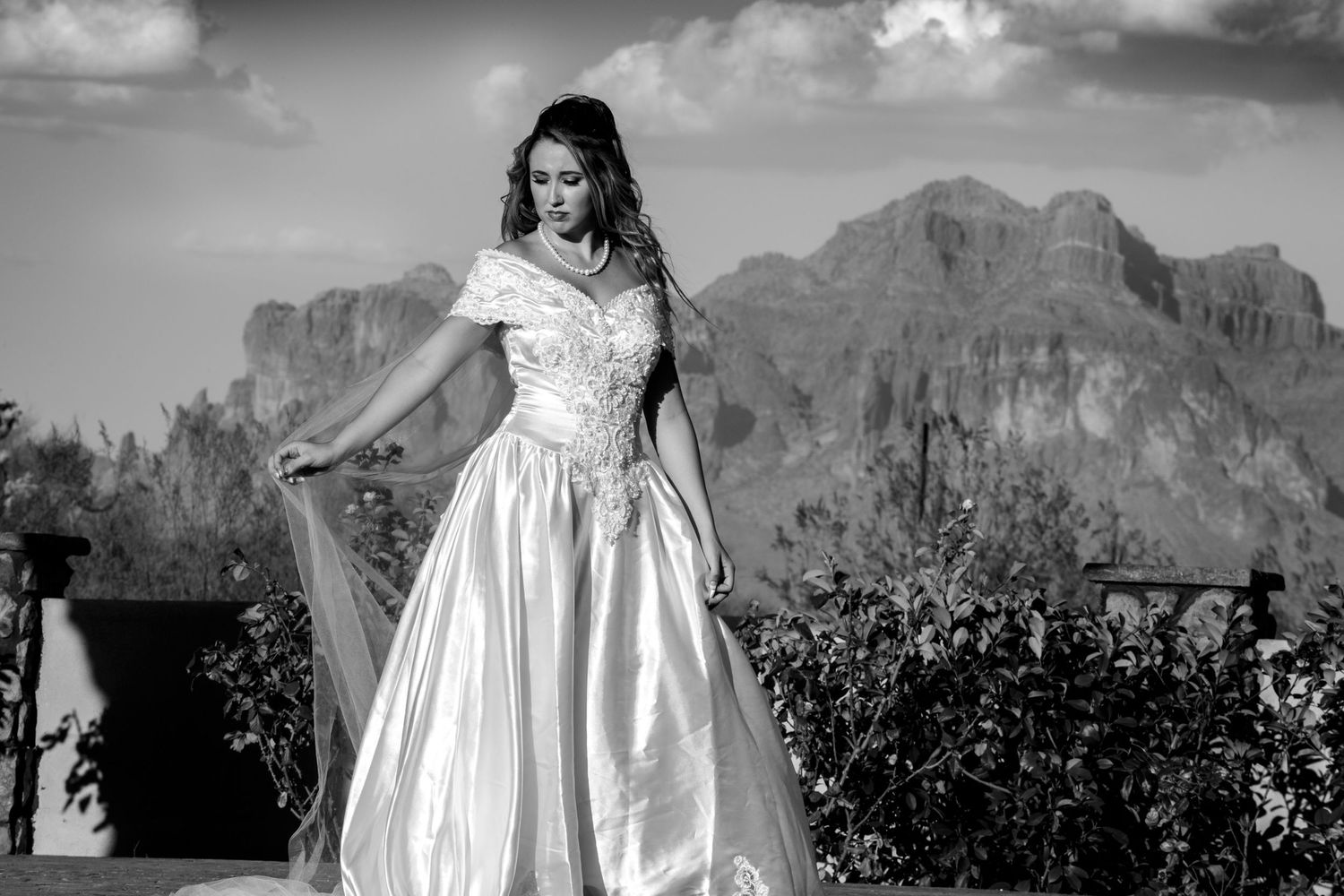 A bride in a white satin wedding dress poses outdoors against a backdrop of dramatic mountain peaks in black and white.