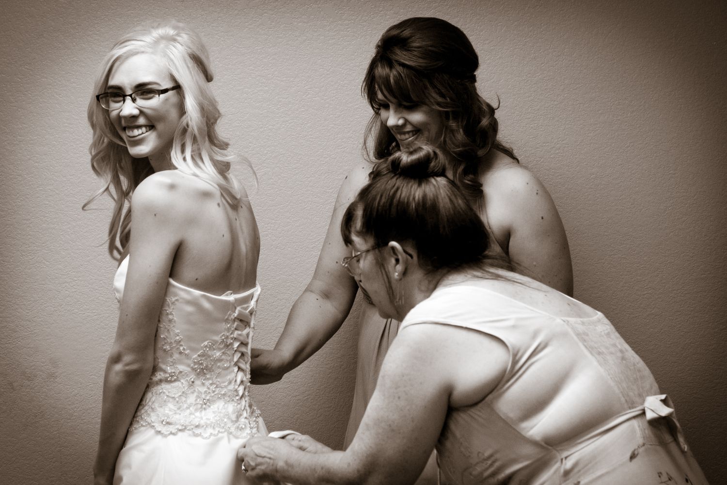 A candid black and white moment captured during bridal preparation showing joyful laughter and final dress adjustments.