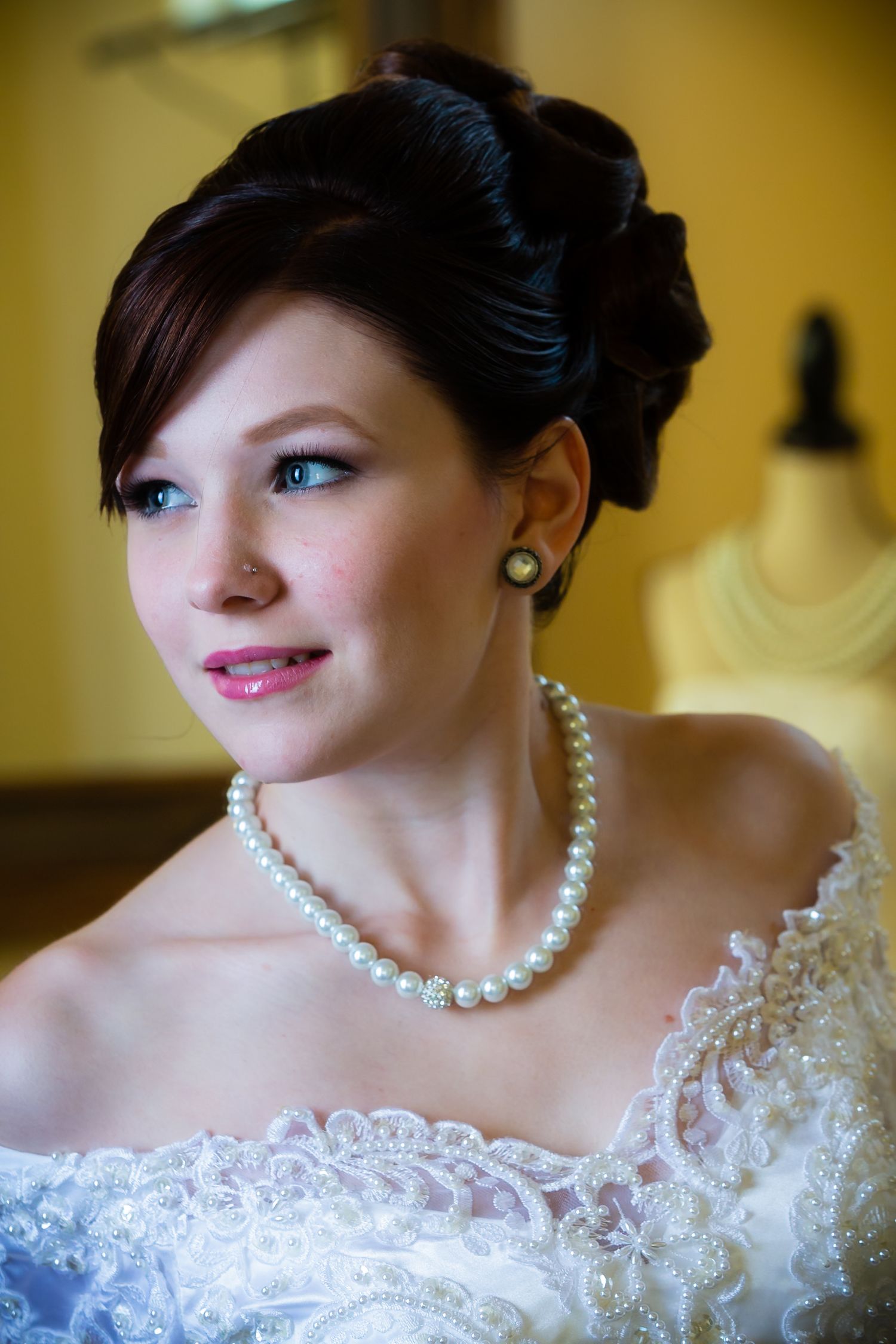 Elegant bridal portrait showing pearl necklace and delicate lace strapless wedding dress against warm lighting.