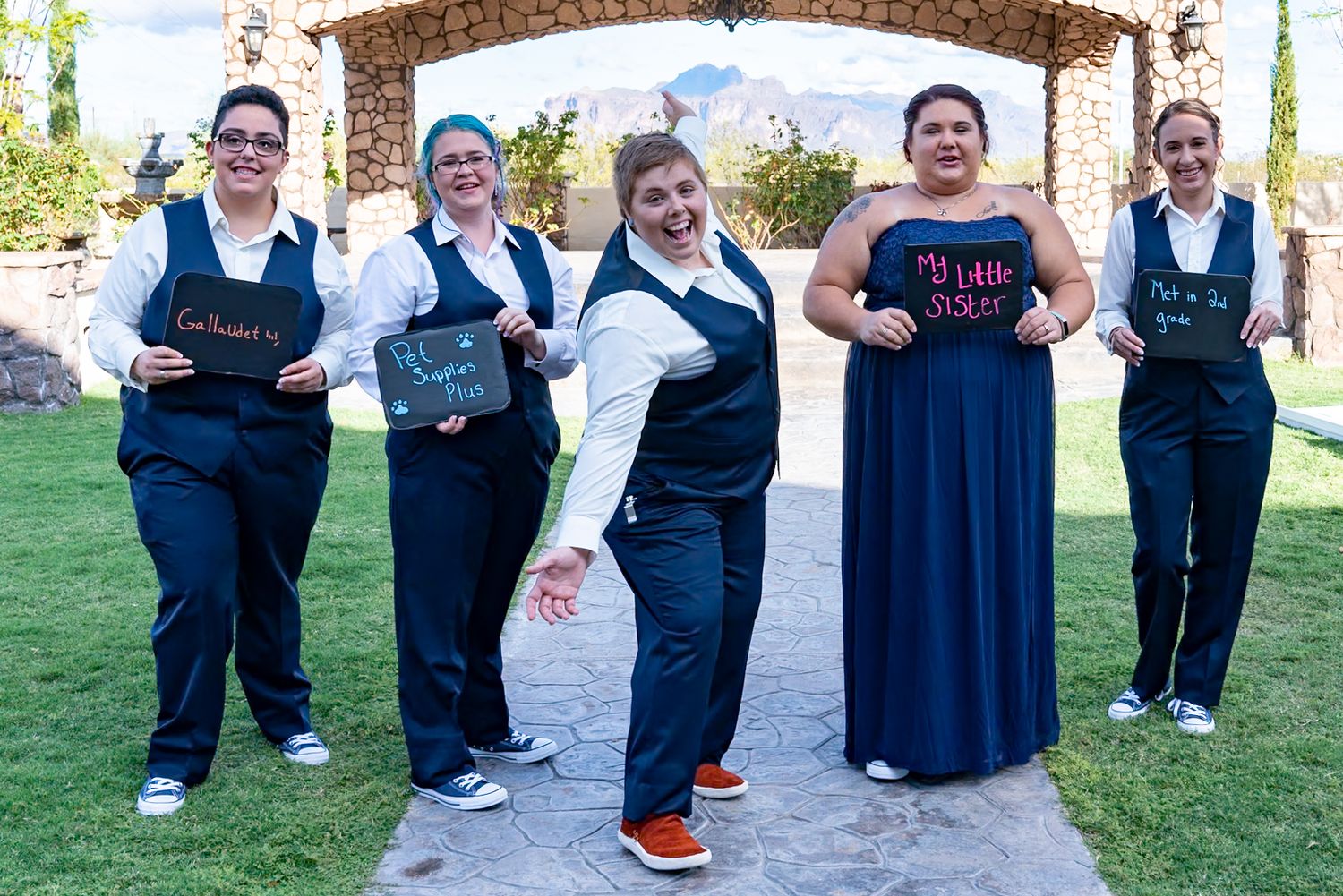 A group in matching navy blue outfits and vests poses together outdoors in front of a garden archway.