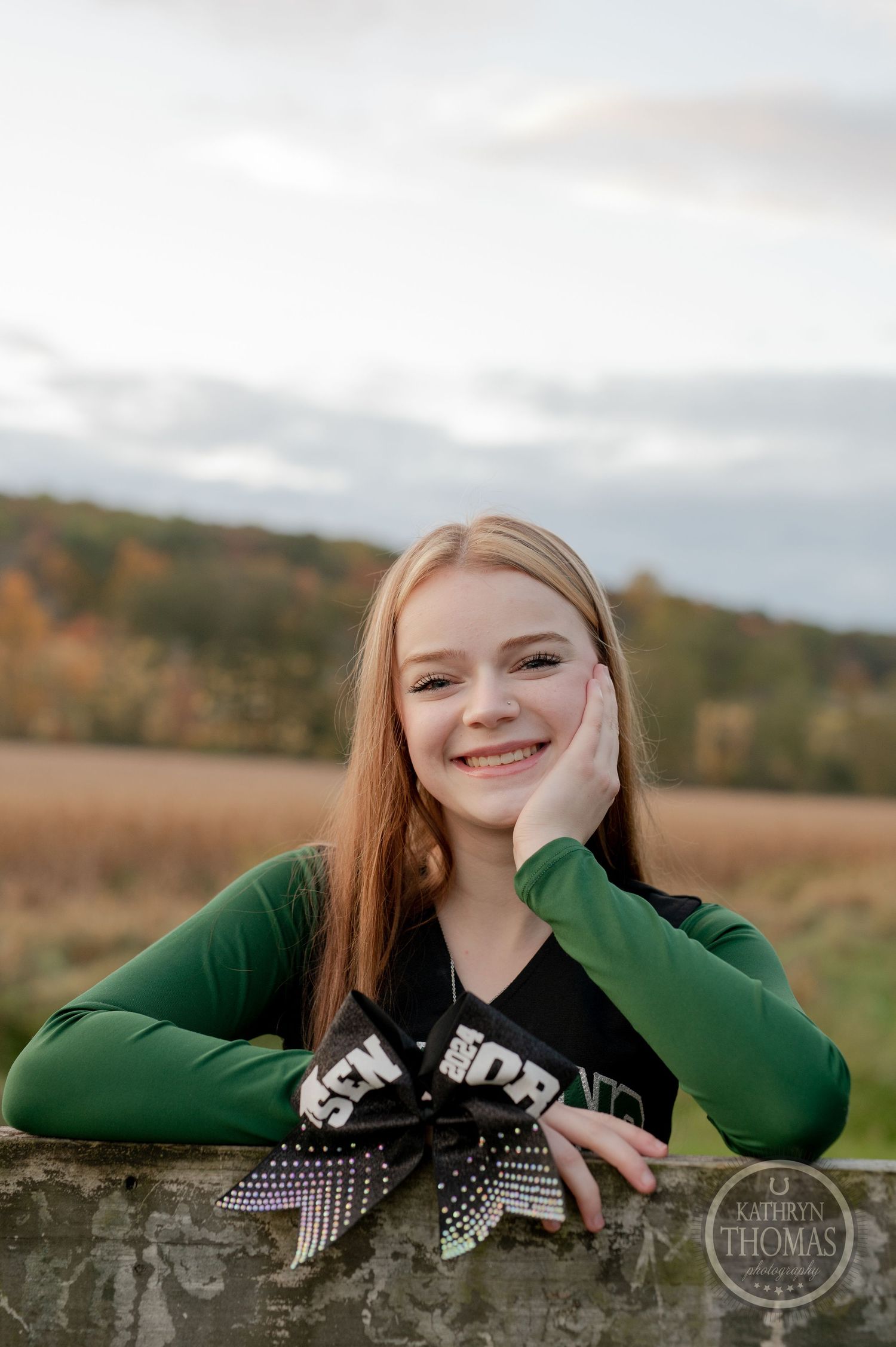 Student in green and black varsity uniform smiles while leaning on wooden fence.