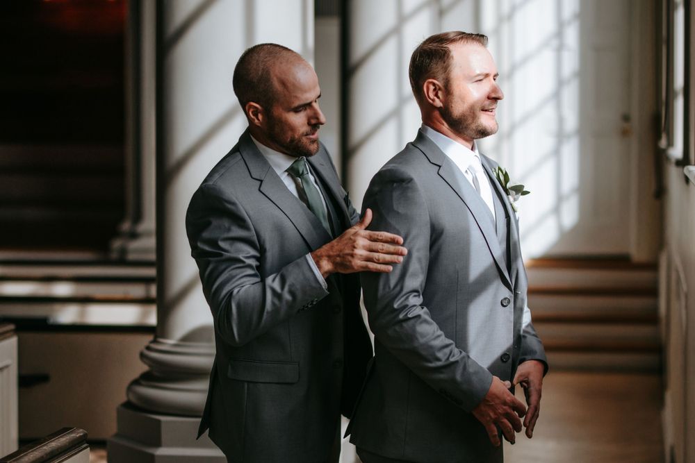 Two men in formal suits stand in an elegant hallway with natural lighting.