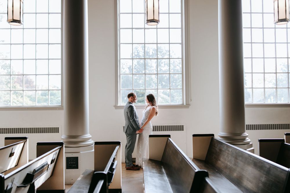 A romantic wedding moment between a couple standing by tall church windows with wooden pews in the foreground.
