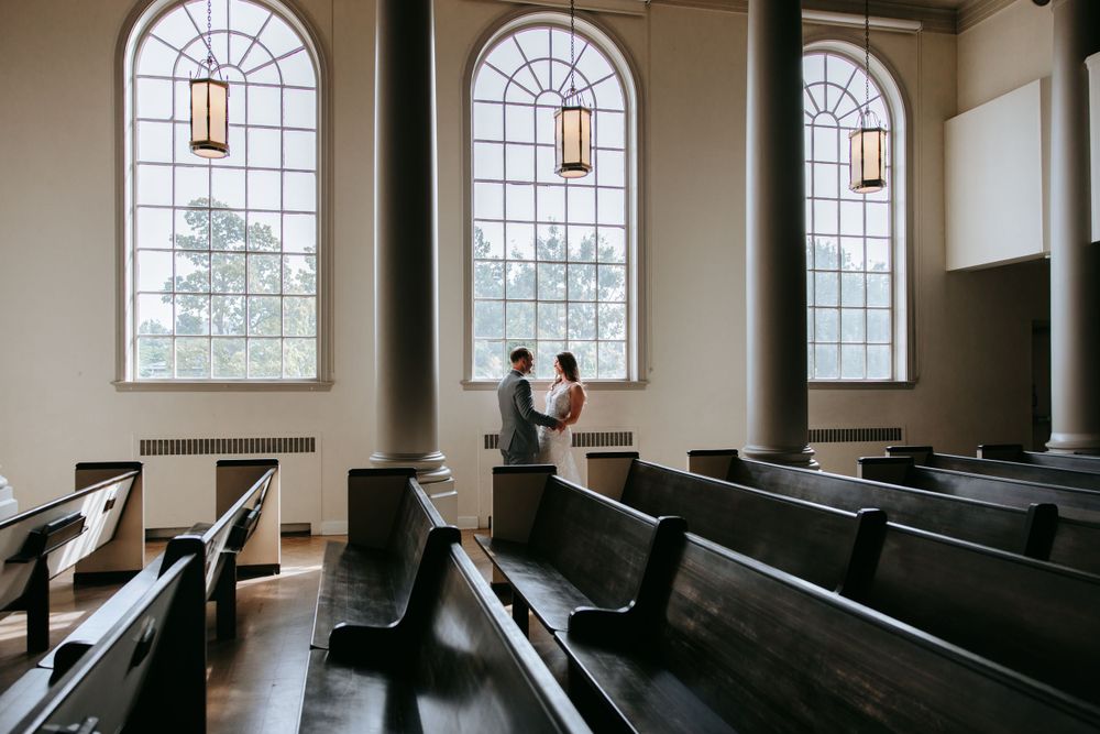 Elegant church interior with arched windows and wooden pews framing a couple in formal attire.