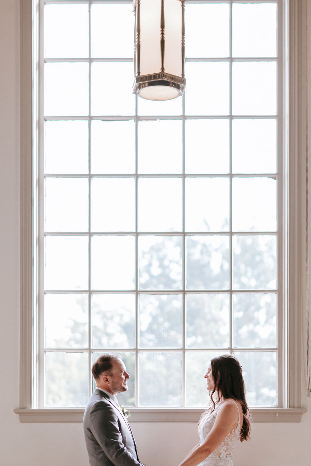 Couple share a tender moment by a tall gridded window with natural light streaming in.
