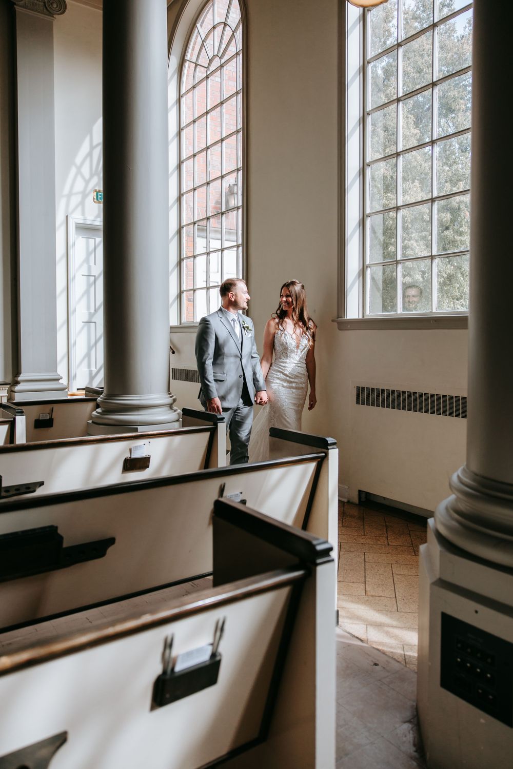 Wedding couple standing among wooden church pews near elegant arched windows.