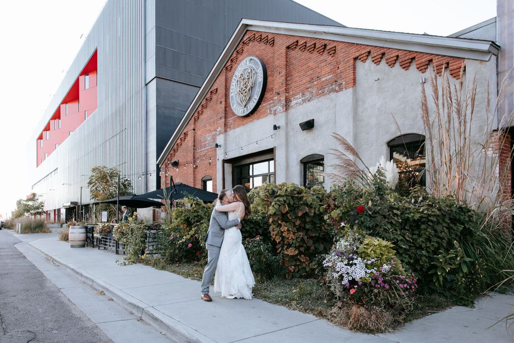 A romantic wedding photo outside a rustic brick building with greenery and decorative lighting.
