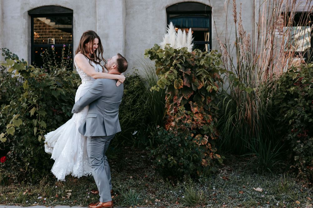 A couple sharing an intimate moment against a backdrop of green shrubs and industrial architecture.