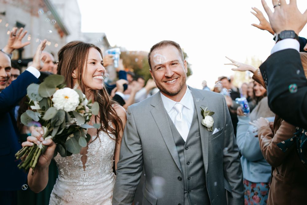 Newlyweds exit their celebration surrounded by joyful guests and white floral bouquets.