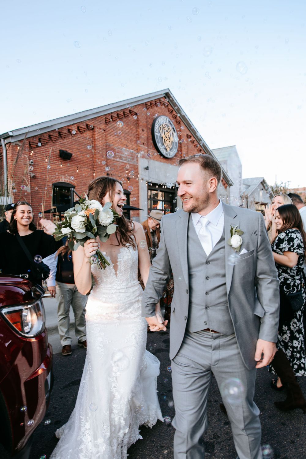 Newlyweds walk together outside a historic brick building with clock tower and celebrating guests.