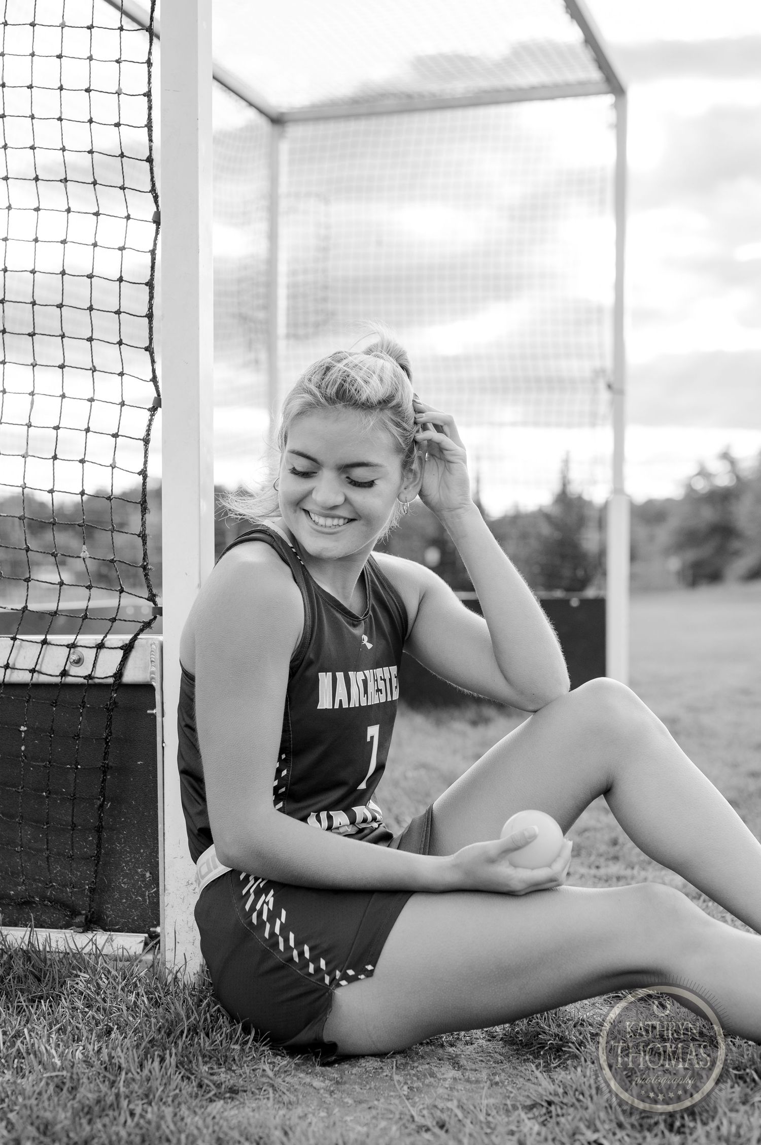 Black and white image shows athlete sitting by field hockey goal in team uniform.