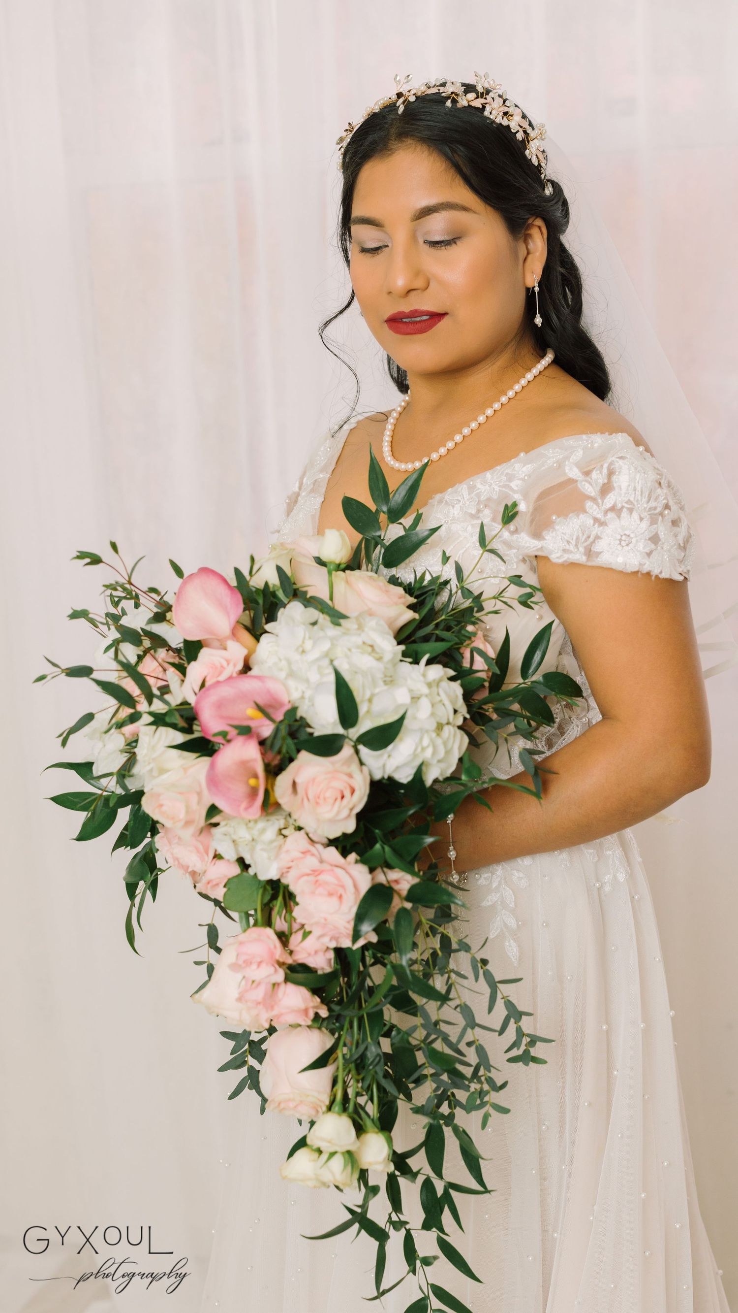 A bride in a white lace dress poses with a cascading pink and white floral bouquet against a light backdrop.