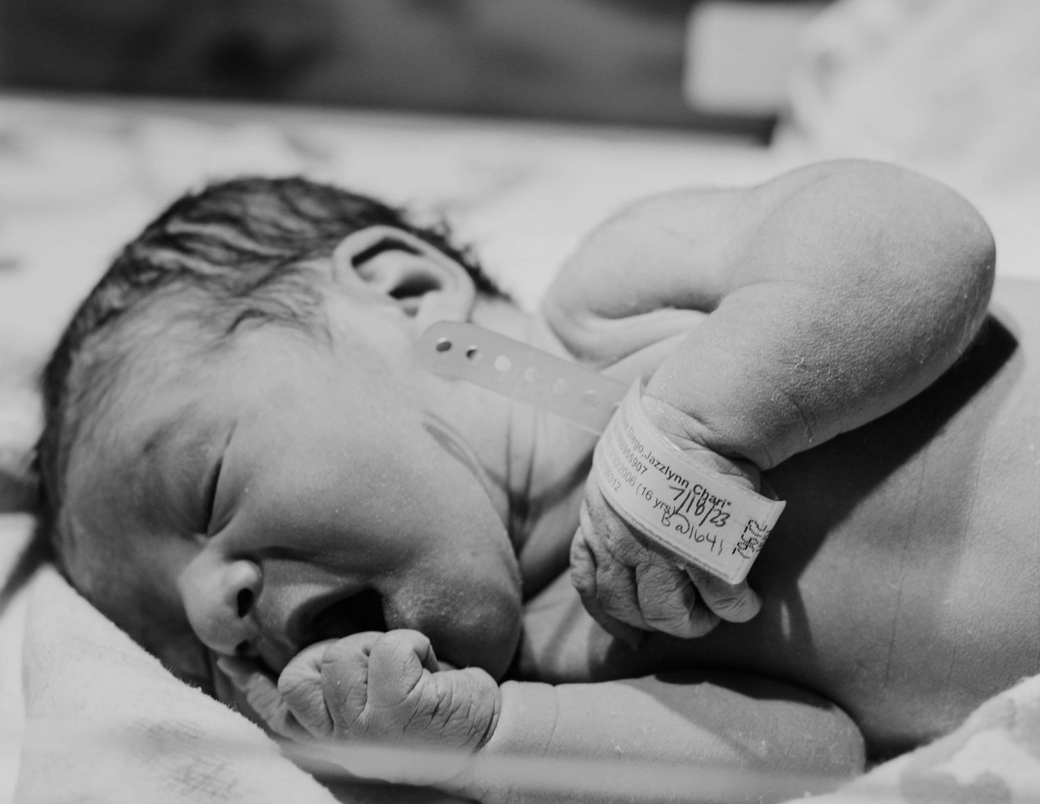 A black and white photograph of a newborn baby lying on a blanket with a hospital ID bracelet on their wrist.