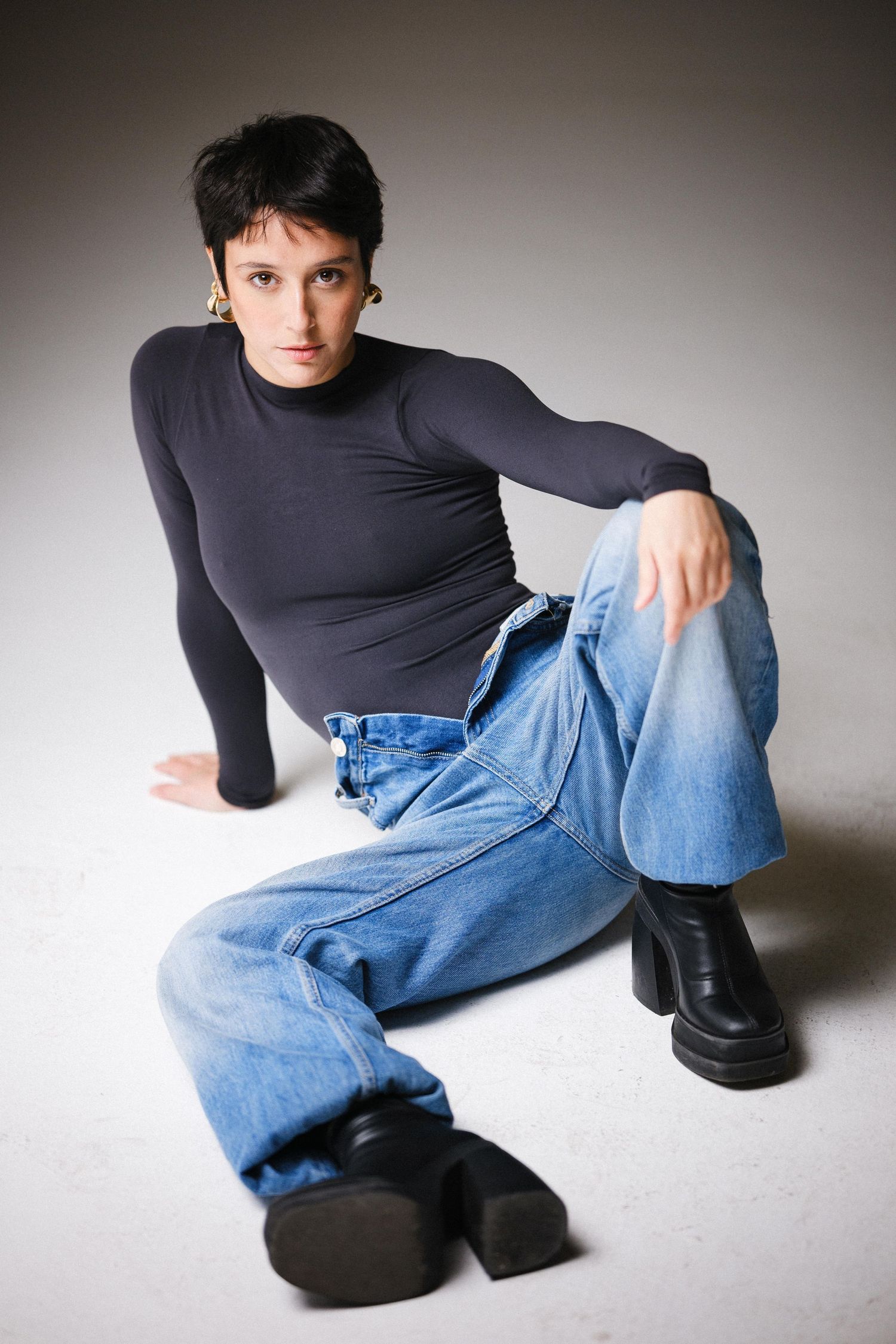 Model in black top and jeans poses on floor with black boots in minimalist studio setting.