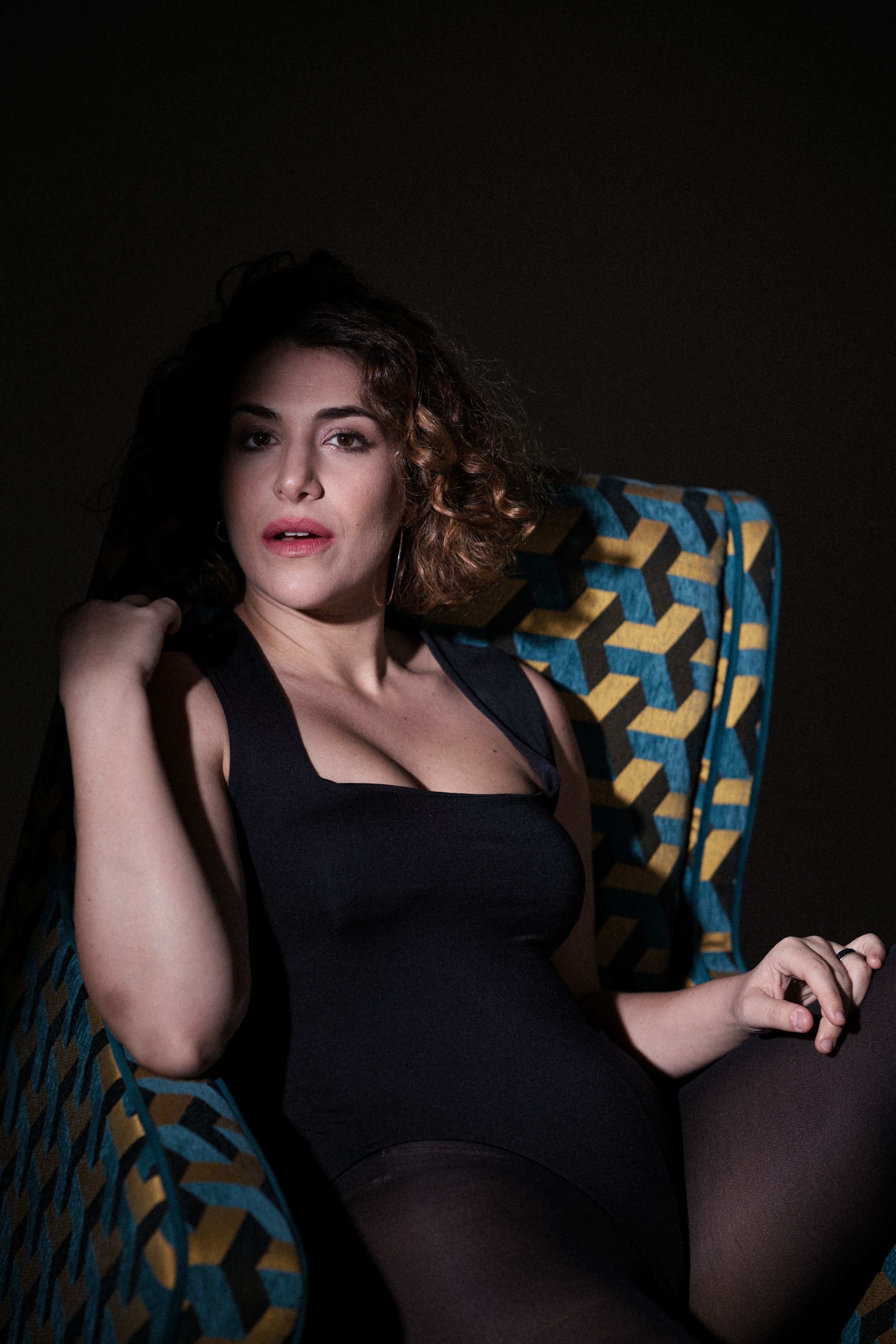 Dramatic boudoir photo of figure in black dress reclining on patterned chair with moody lighting.