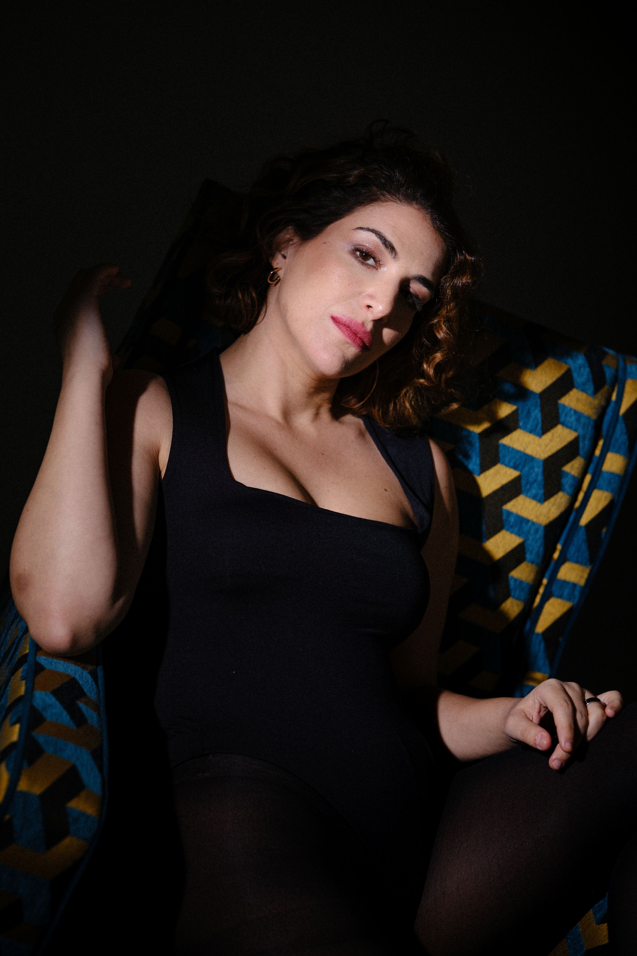 Artistic portrait with dark shadows showing person in black dress against geometric patterned furniture.