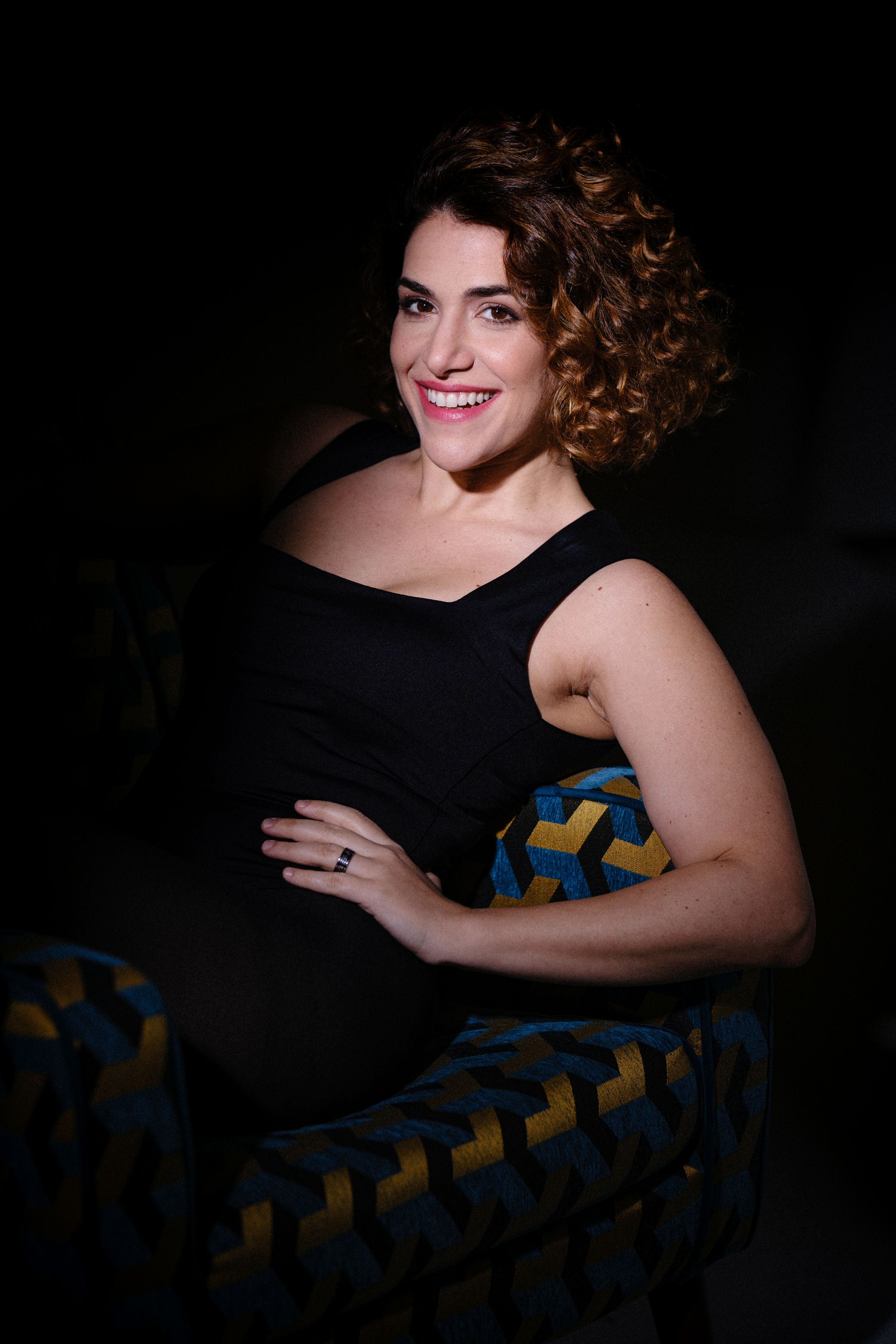Portrait of a smiling performer in a black sleeveless dress seated on a patterned chair against dark background.