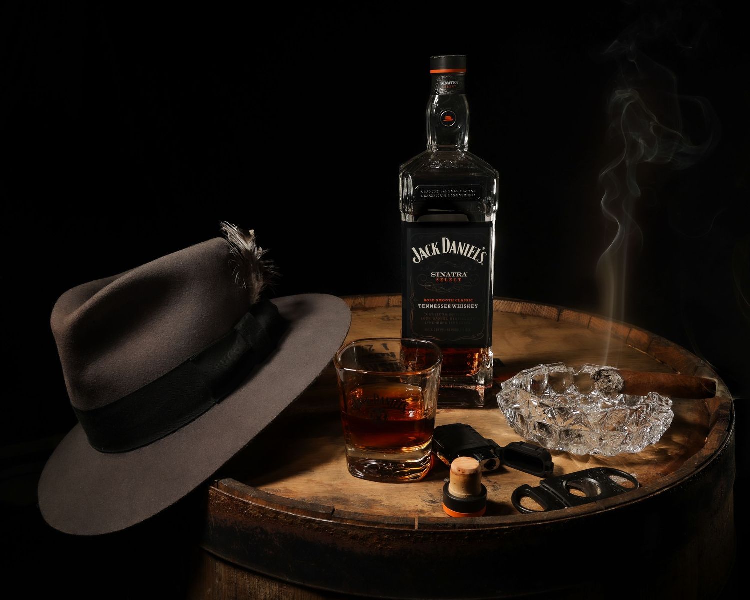 A dark hat, Jack Daniel's whiskey bottle, and glass with smoking cigar on wooden barrel with moody dark lighting.