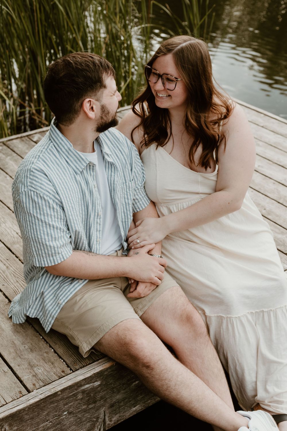 A couple sits closely on a dock by the water, smiling and holding hands in summer.