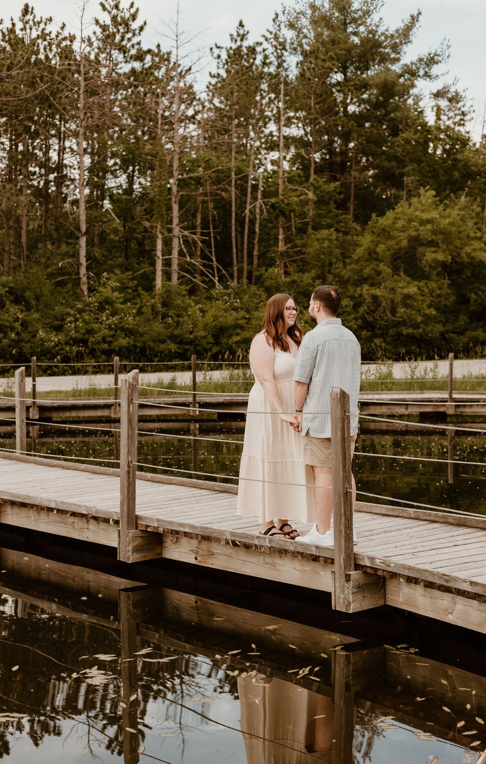 A couple holds hands on a wooden dock surrounded by lush green trees and calm water.