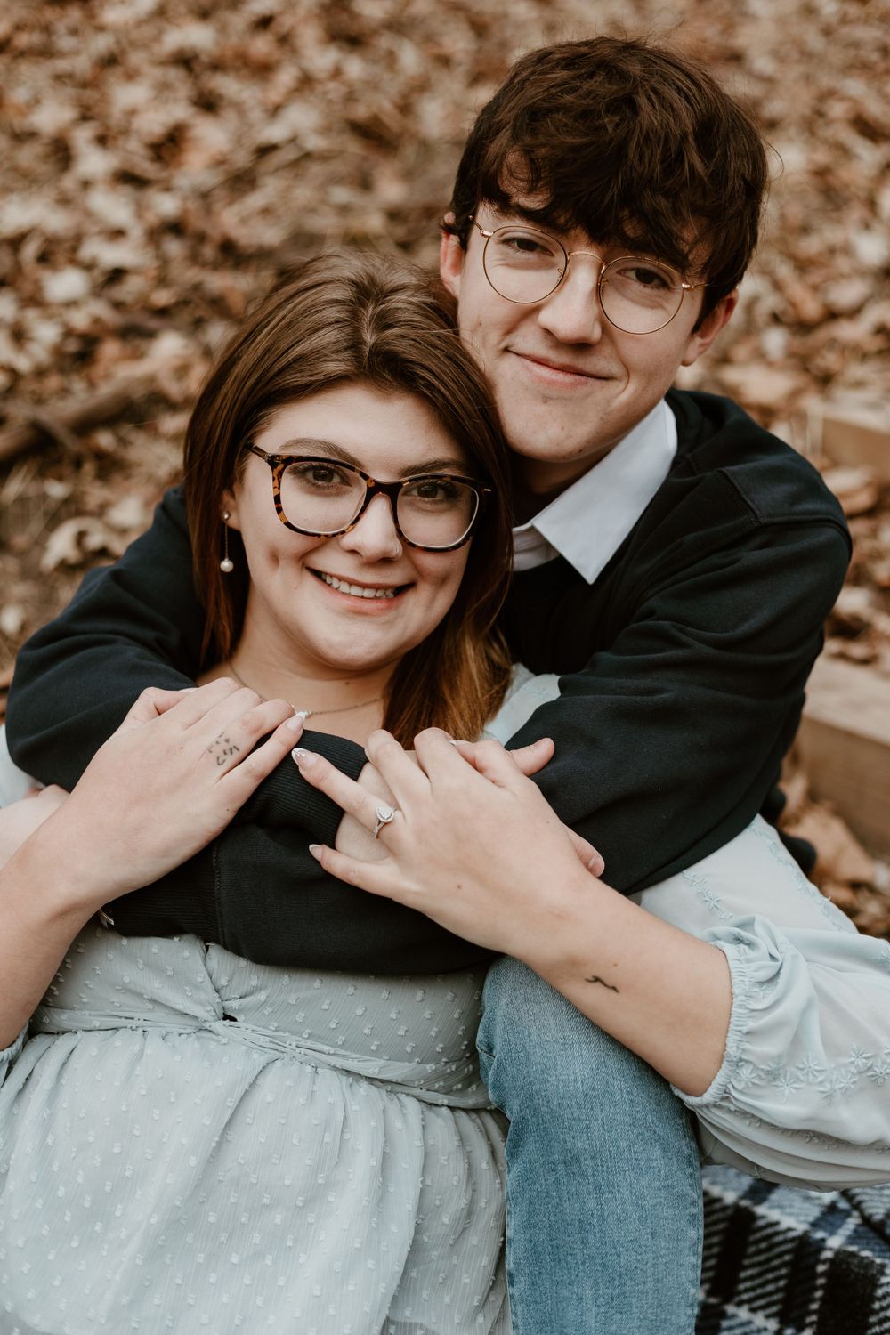 Engaged couple wearing glasses embrace warmly, showing off engagement ring among autumn leaves.