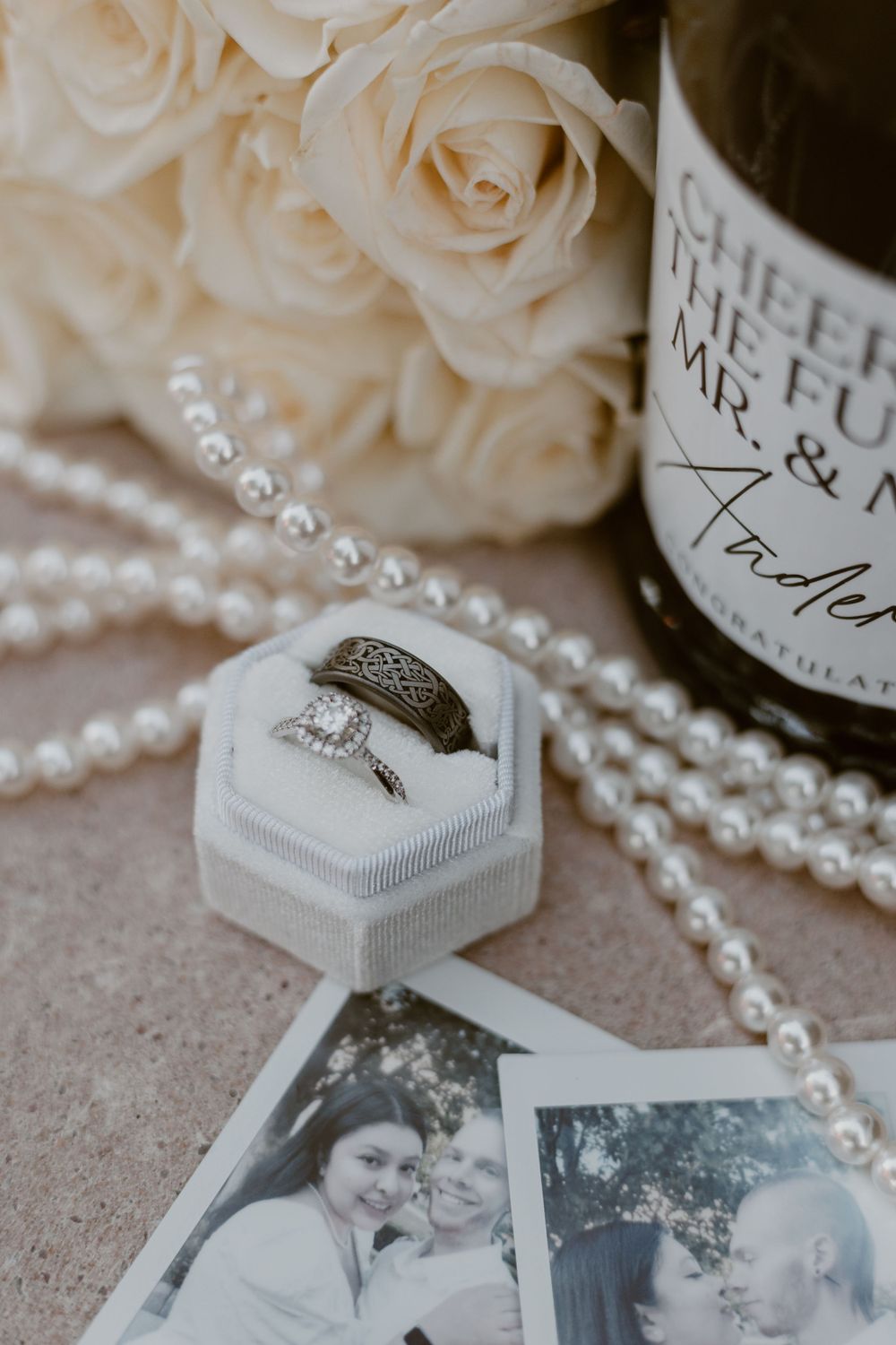 Wedding rings in a white box surrounded by roses, pearls, photos, and a champagne bottle.