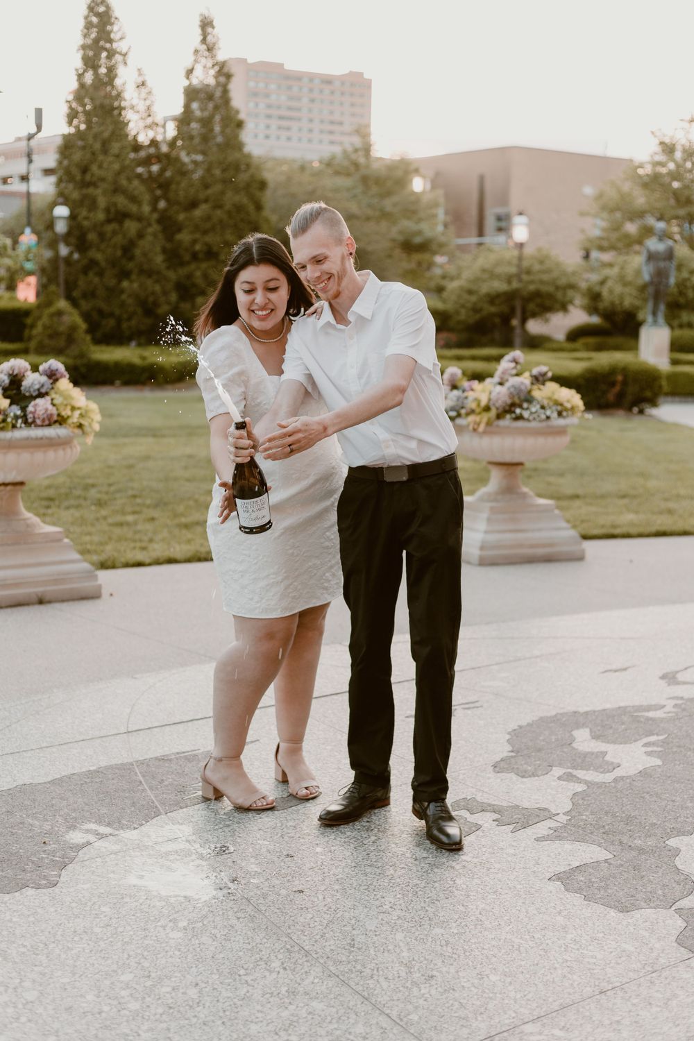 Newlyweds celebrate outdoors popping champagne bottle in wedding attire at sunset.