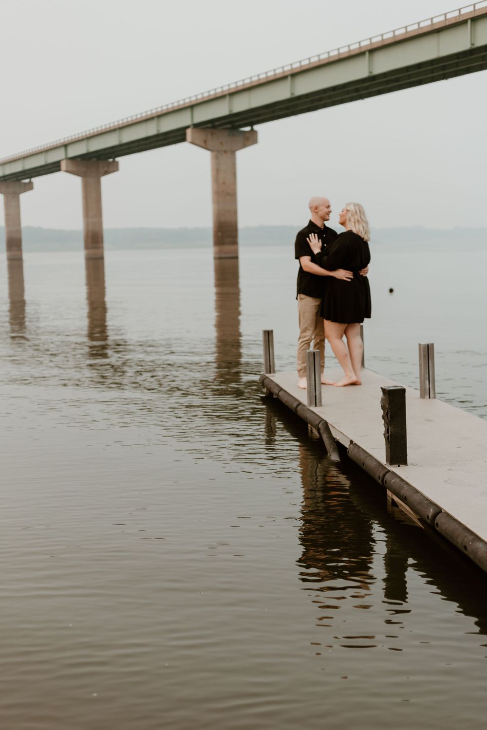 Couple embracing on a wooden dock over calm water with a large bridge in the background.