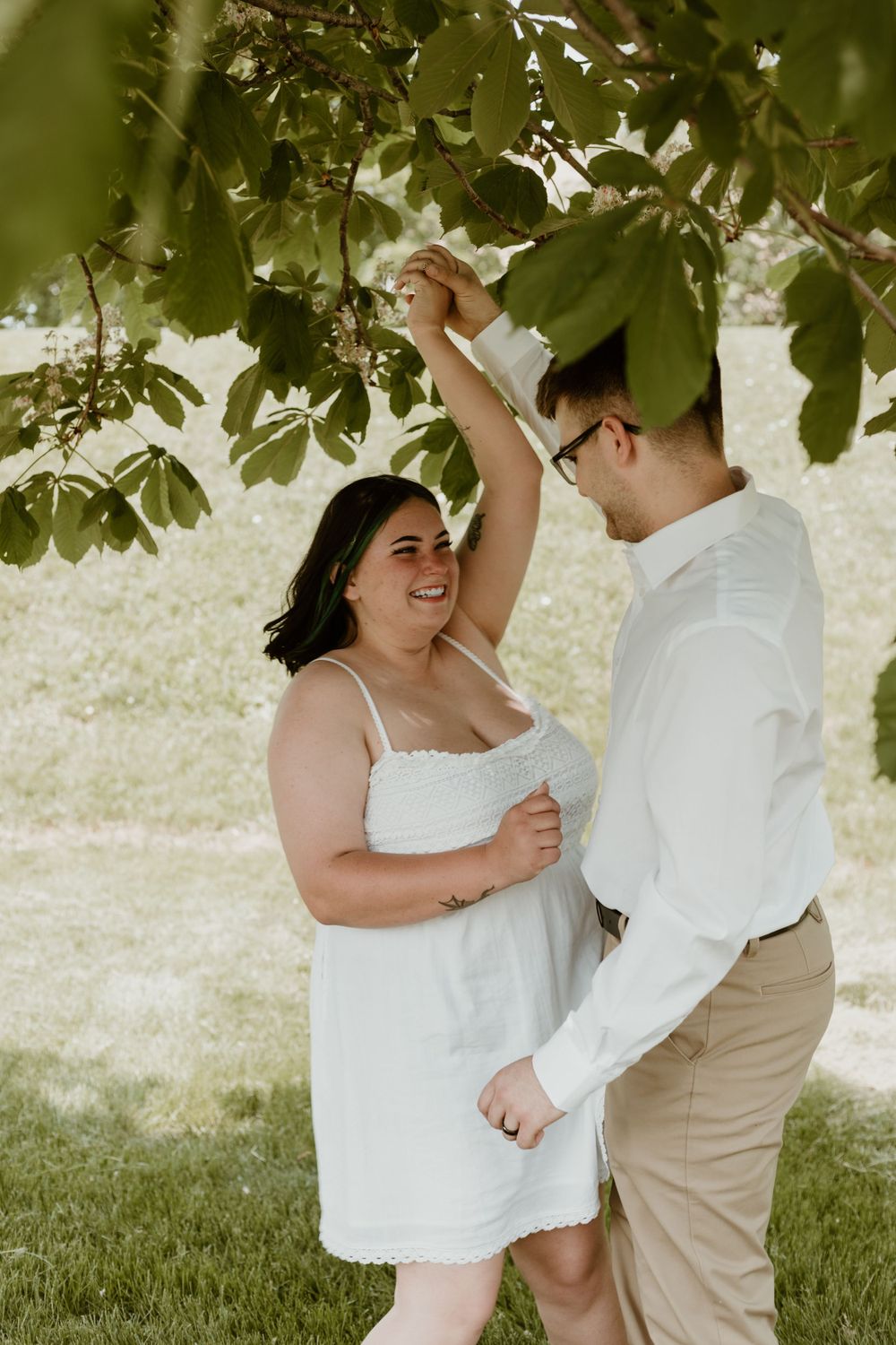 Joyful couple dancing under a tree in a sunny park during an outdoor engagement session.