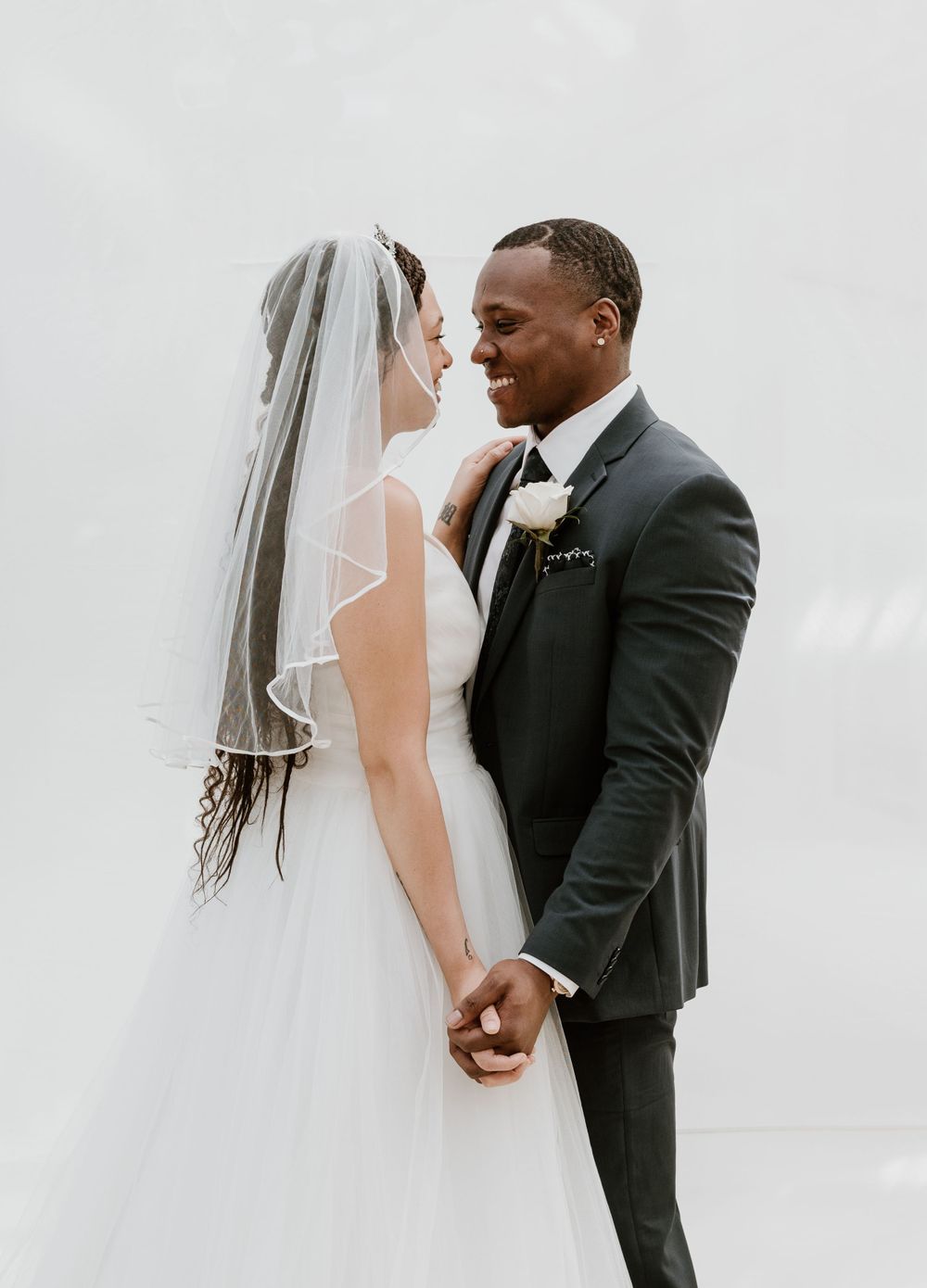 Bride and groom in black suit share intimate moment holding hands against white background.