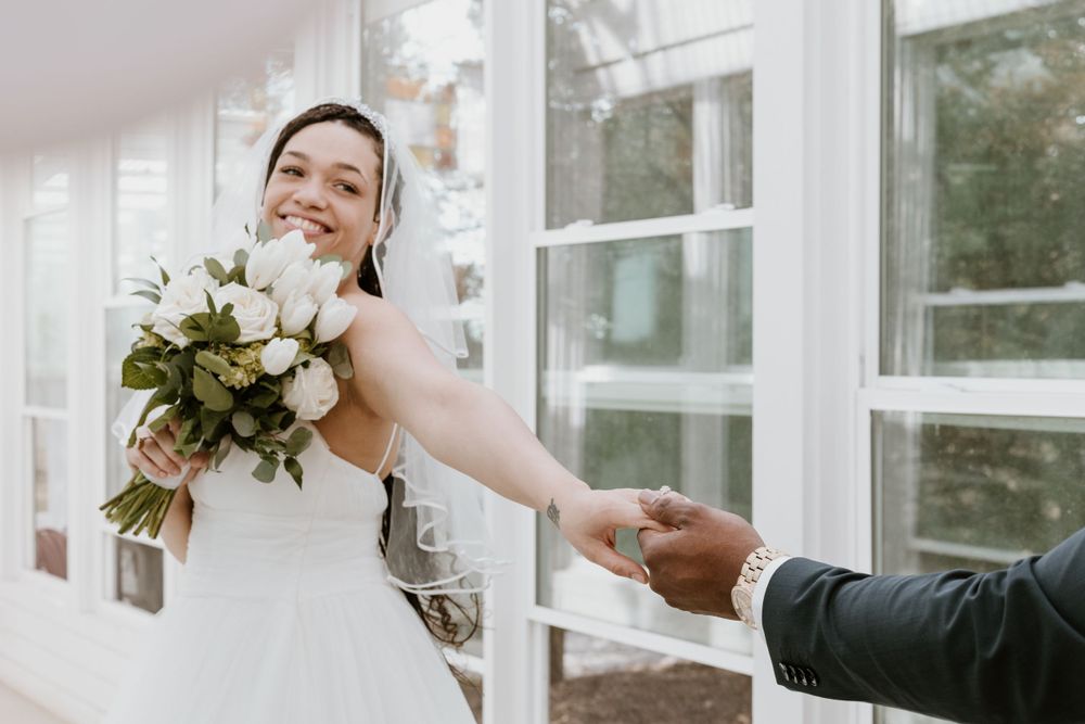 Smiling bride in white dress holding bouquet of white flowers while reaching out to hold hands with groom.