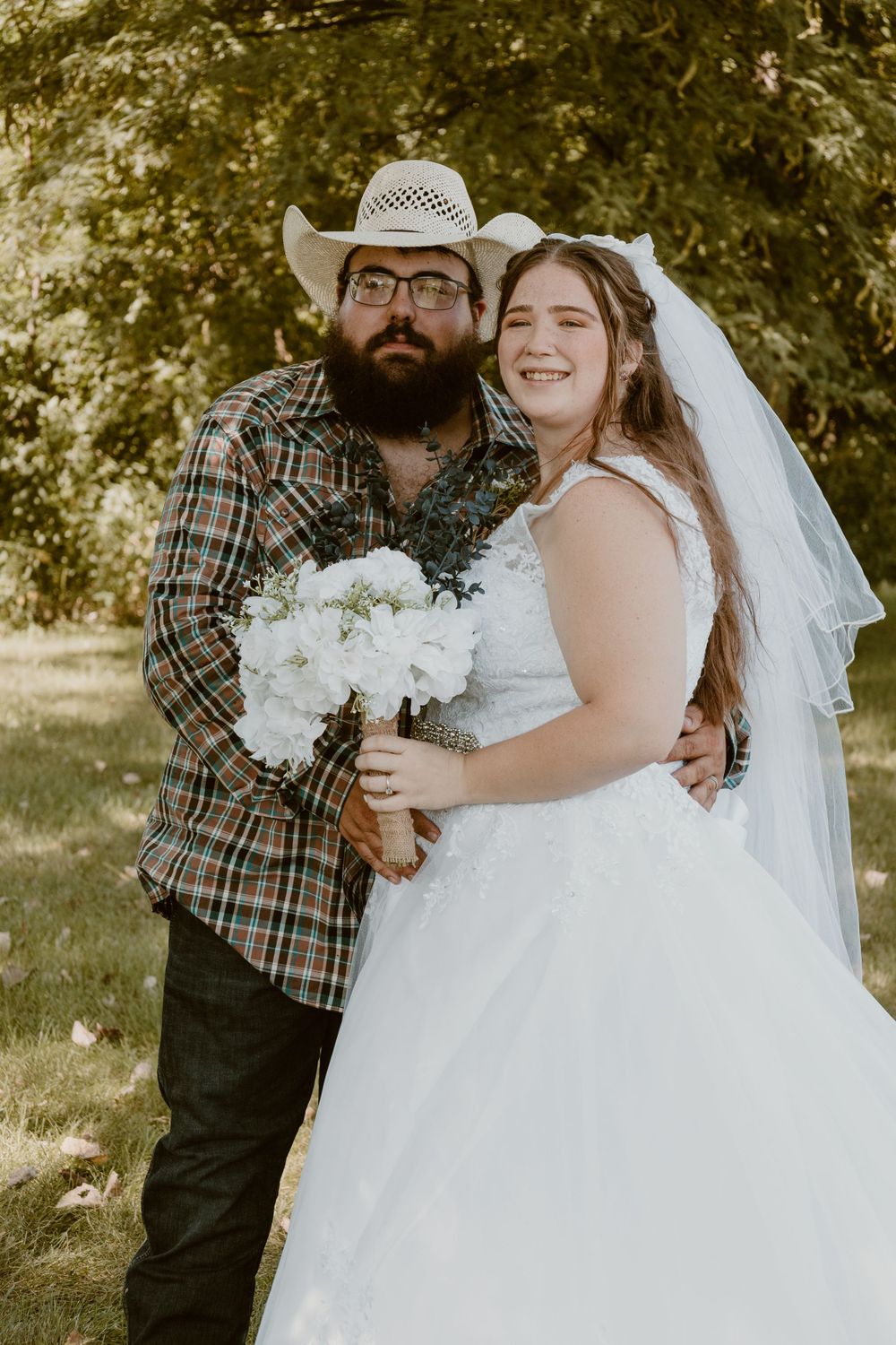 Rustic wedding couple poses outdoors, groom in cowboy hat holding white bouquet with veiled bride.