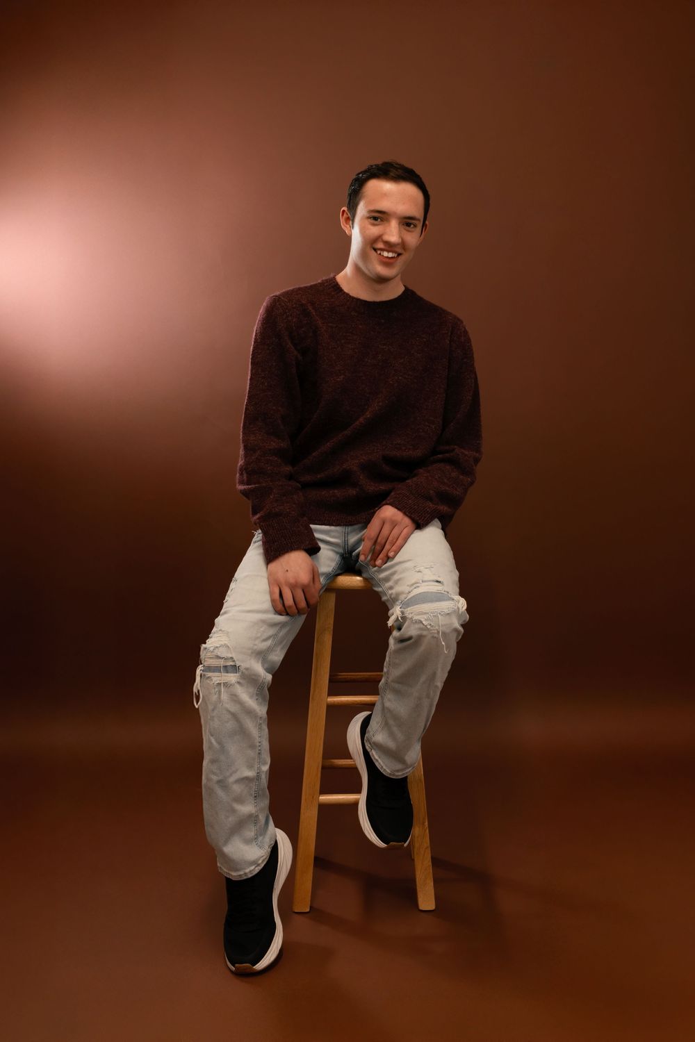 Person in dark brown sweater and distressed jeans sits casually on wooden stool against warm brown studio backdrop.