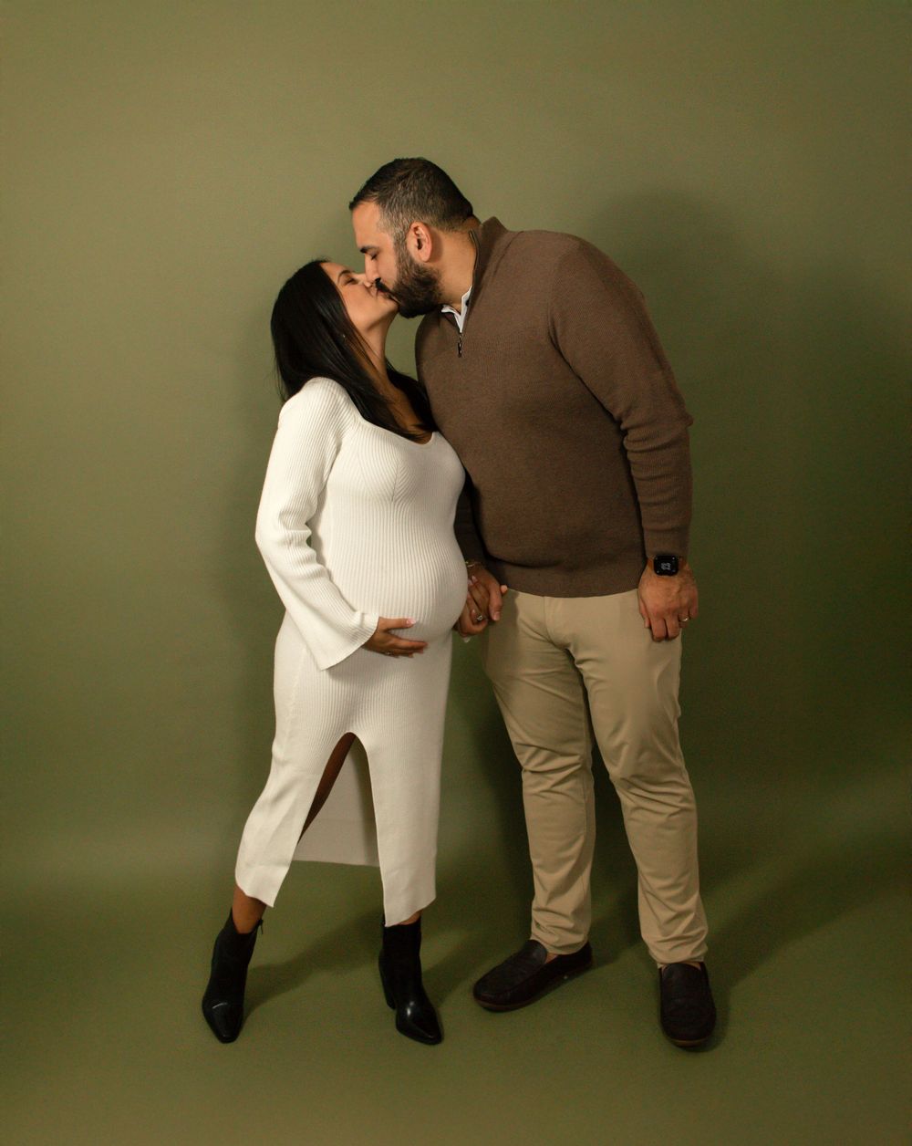 A couple shares an intimate moment against a green studio backdrop, with the woman wearing white and the man in brown.