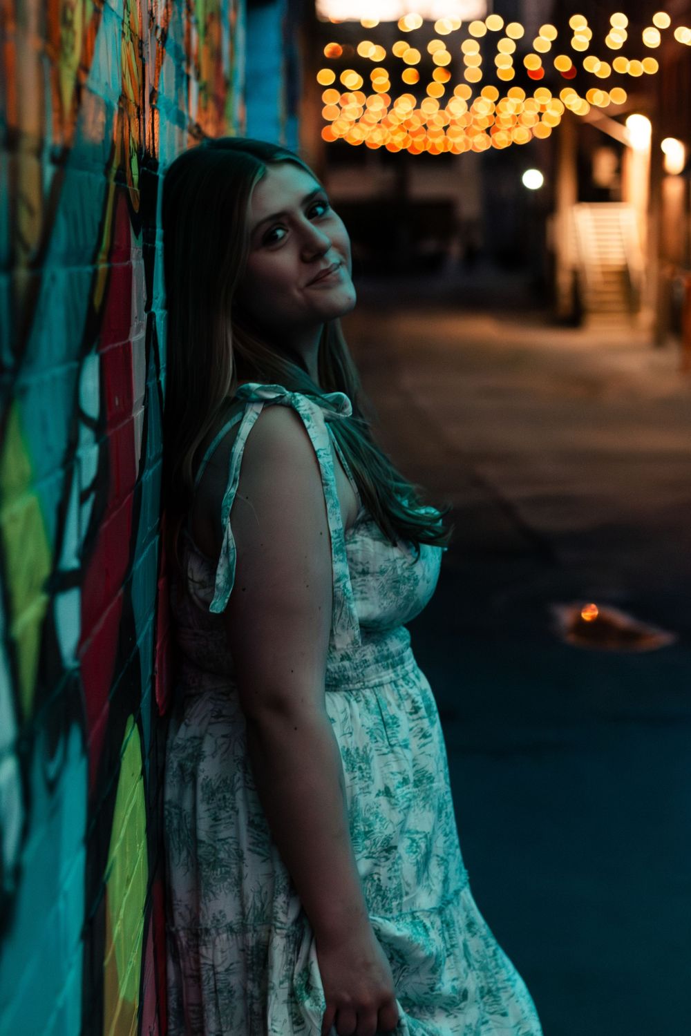 A person in a light dress leans against a wall at night with bokeh lights glowing in the background.