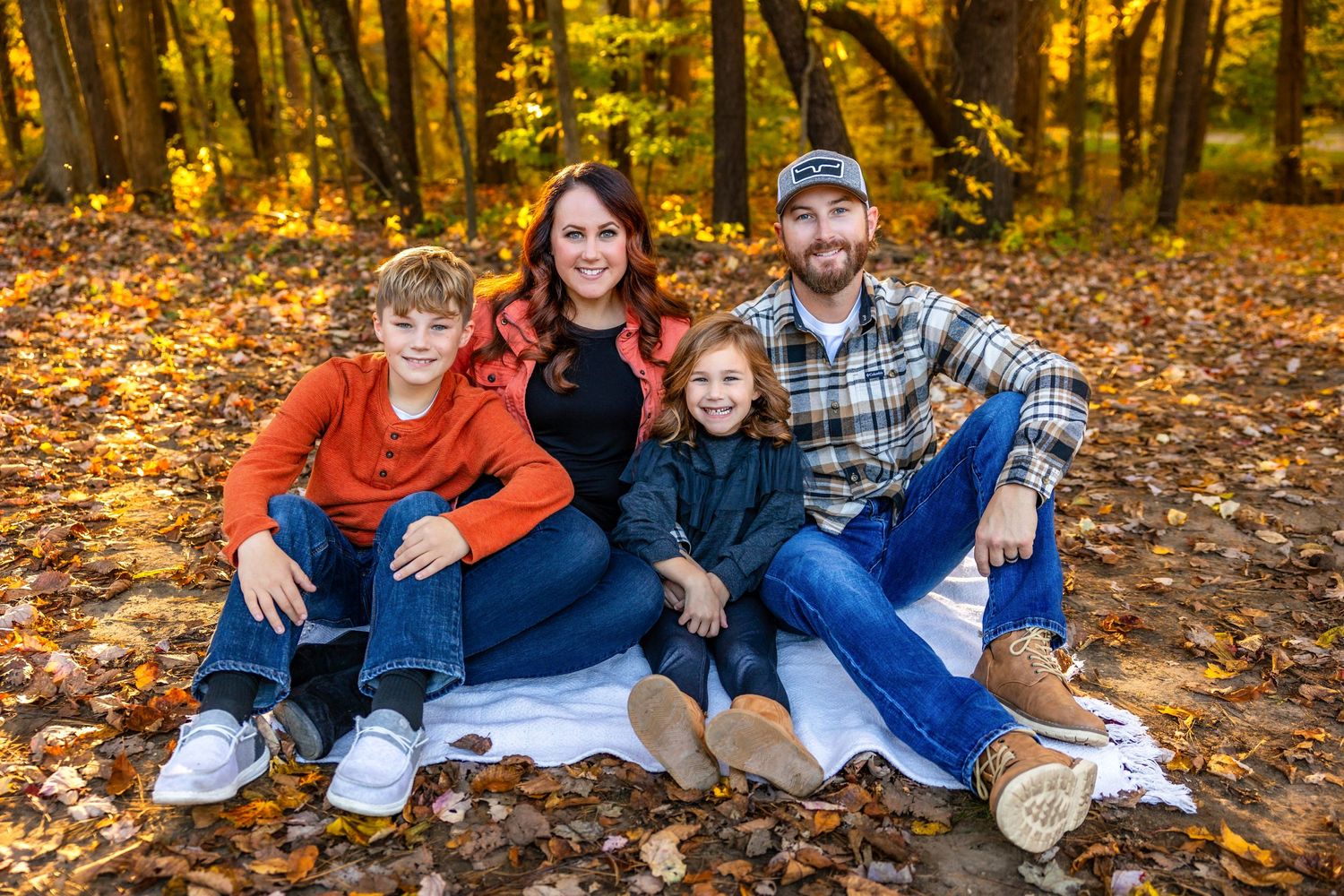 A group sits together on a blanket surrounded by golden fall foliage in a sunlit forest setting.