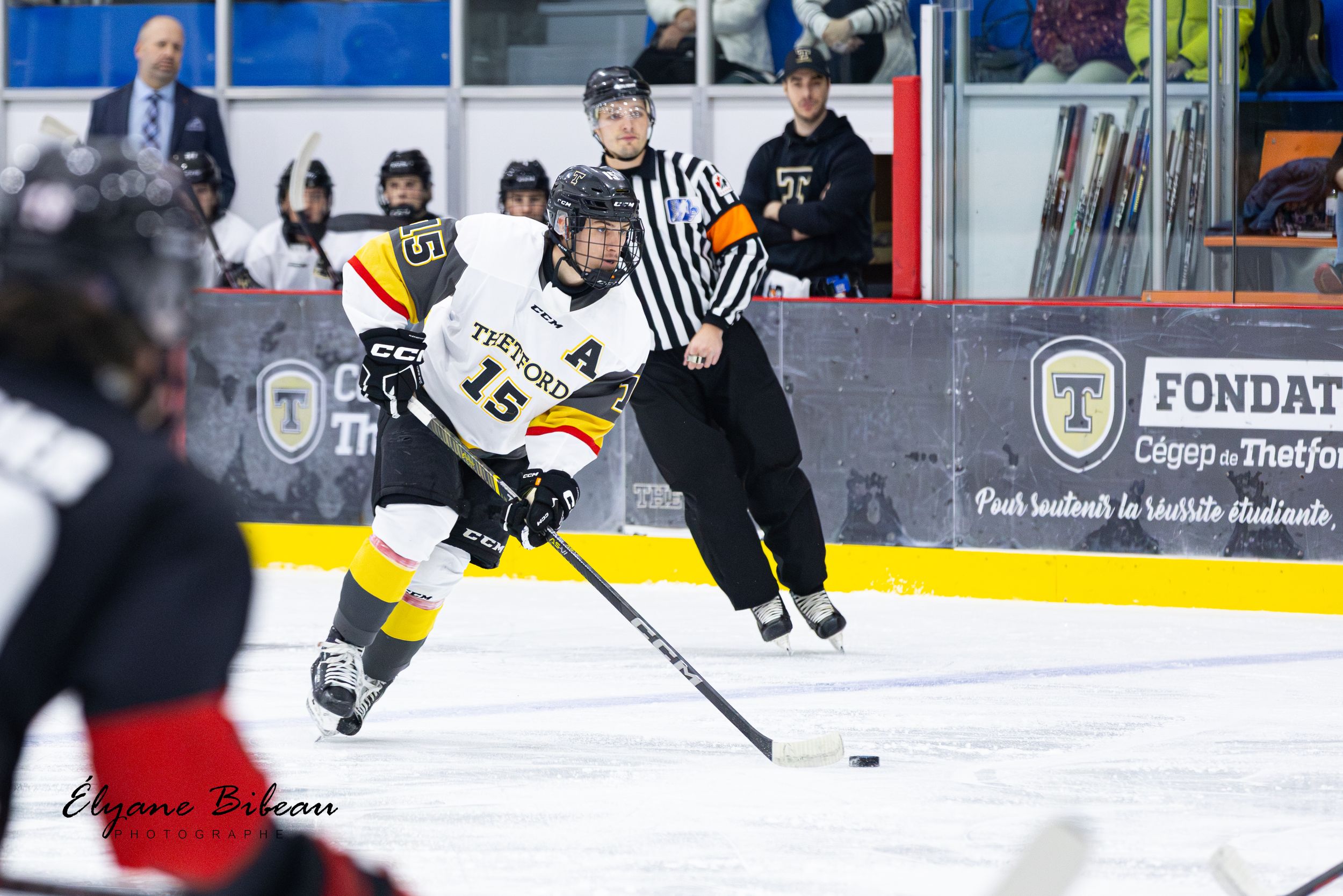 Ice hockey player in yellow jersey controls puck while referee observes nearby during game.