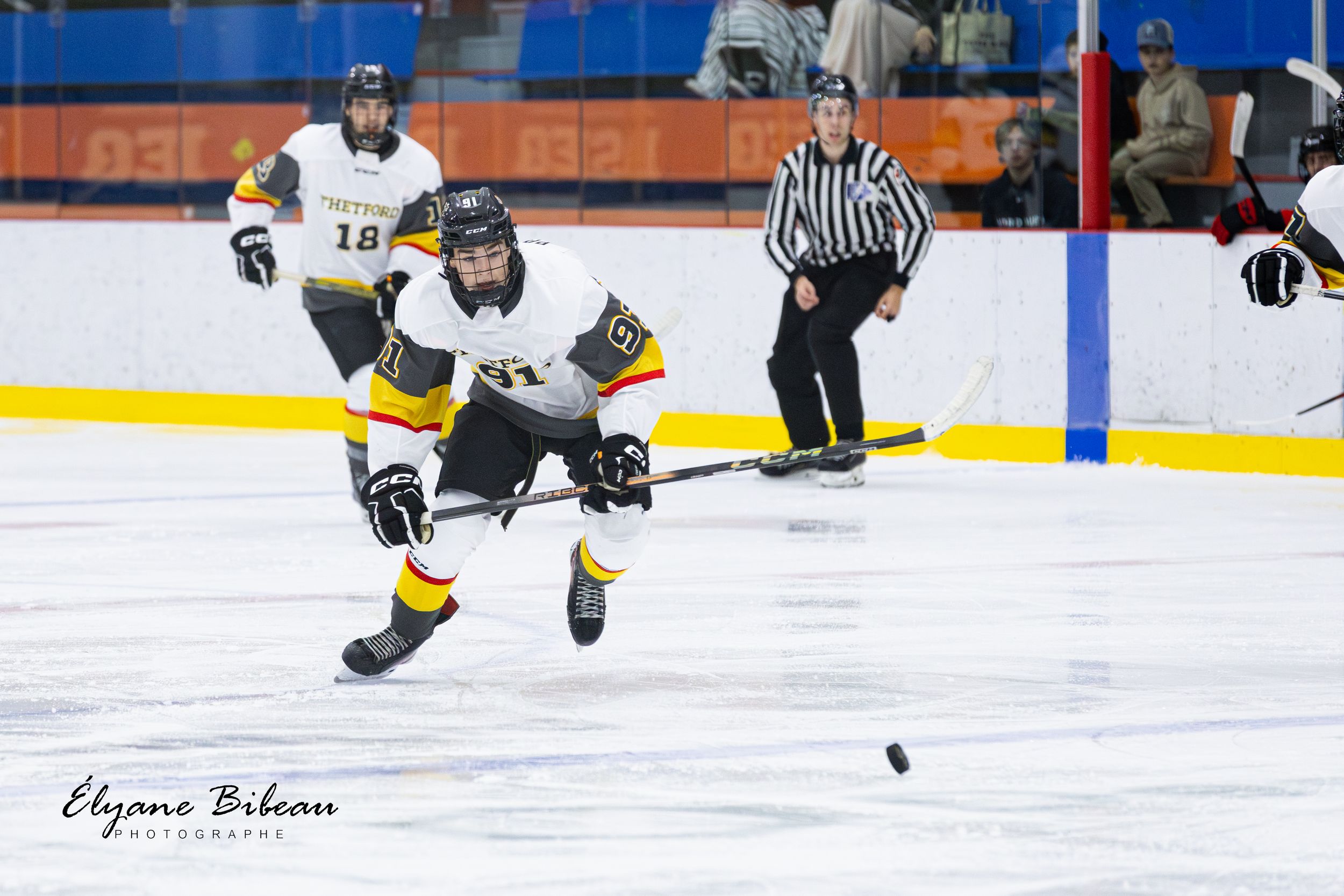 Hockey player in white jersey skates forward chasing after loose puck on ice.