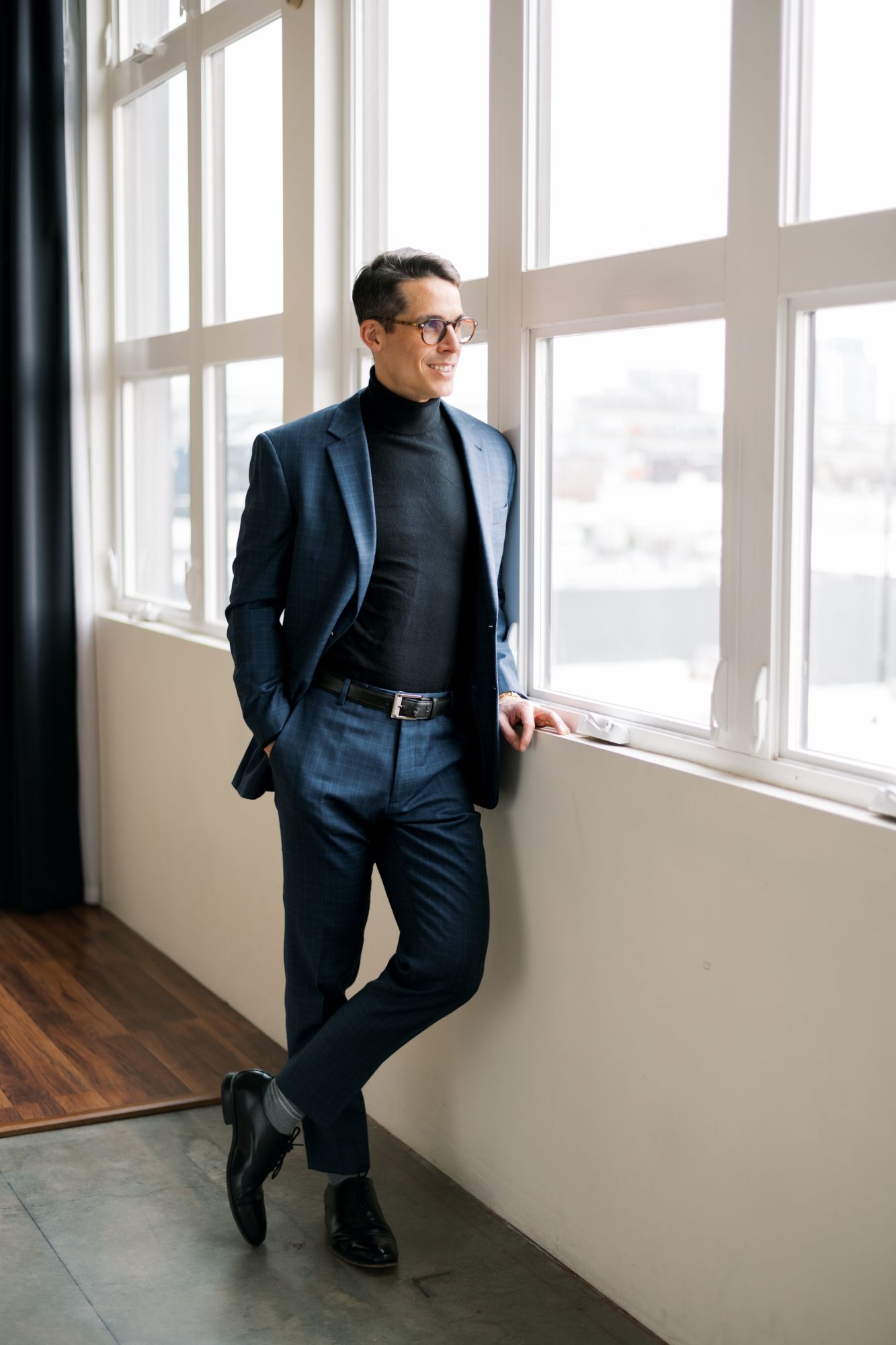 A person wearing a plaid brown blazer, black turtleneck, brown pants and black heels poses on a stool against a gray wall.