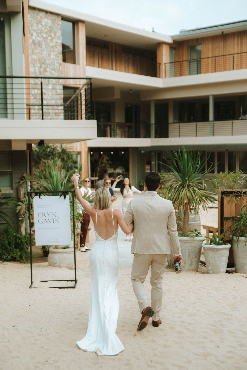 A couple in wedding attire walks through a modern courtyard with stone walls and tropical plants at an outdoor venue.