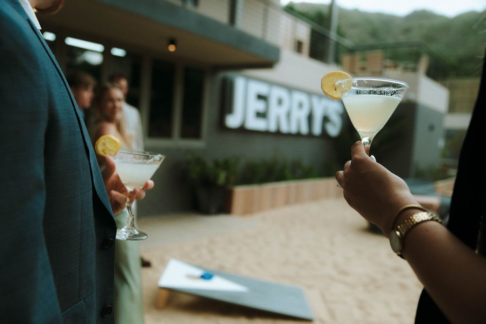 A hand holds a cocktail glass filled with a golden drink garnished with citrus against a dark background.
