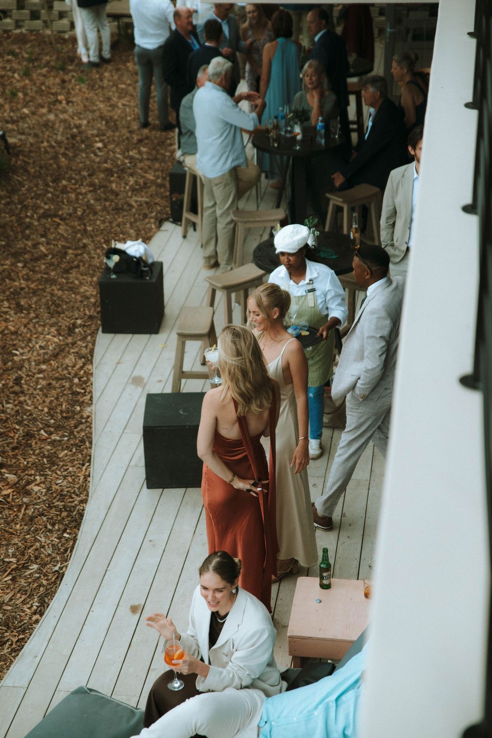 Wedding guests mingle and socialize on an outdoor wooden deck during a reception gathering.