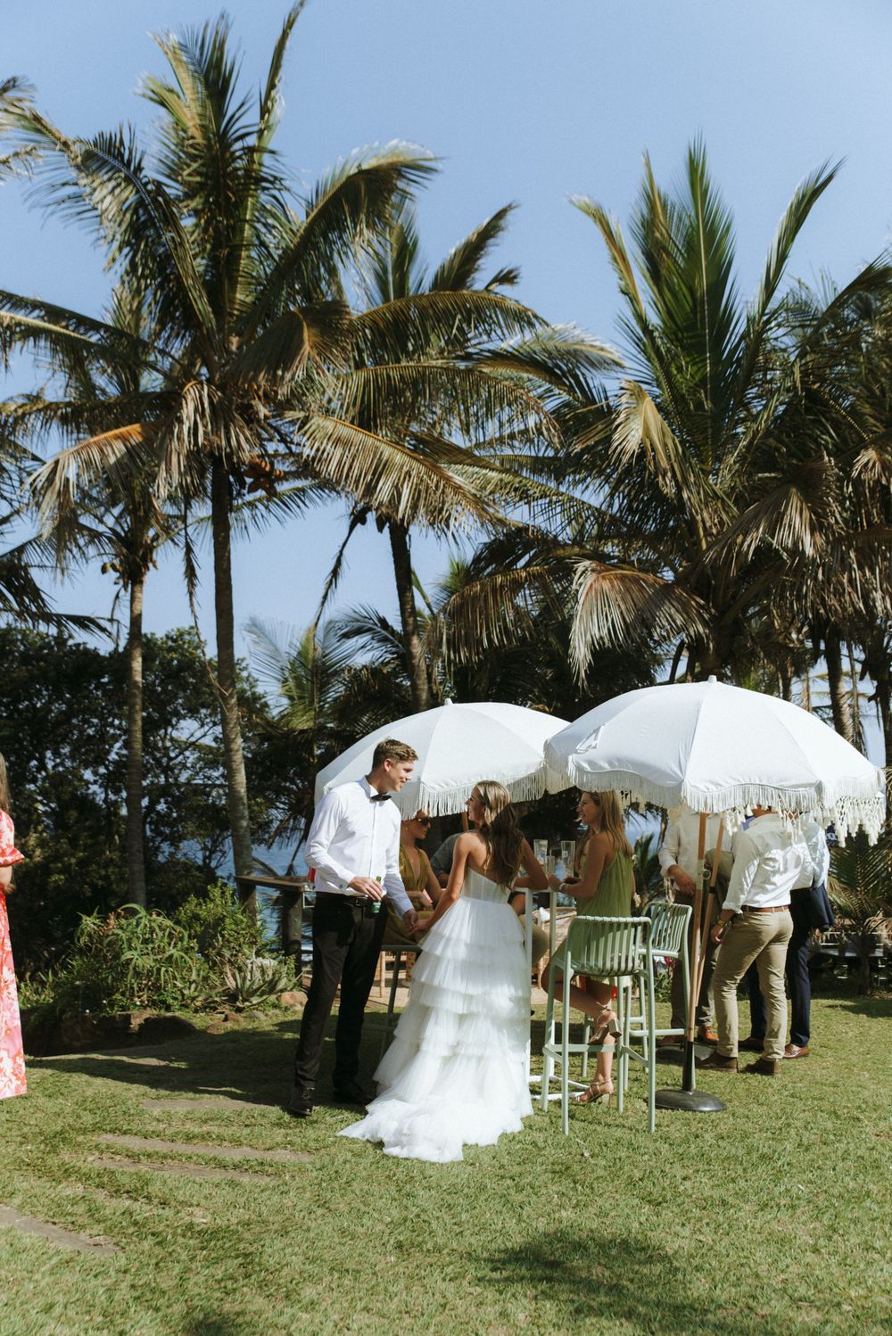 A tropical outdoor wedding ceremony setup with white umbrellas and palm trees swaying in the breeze on a grassy hillside.