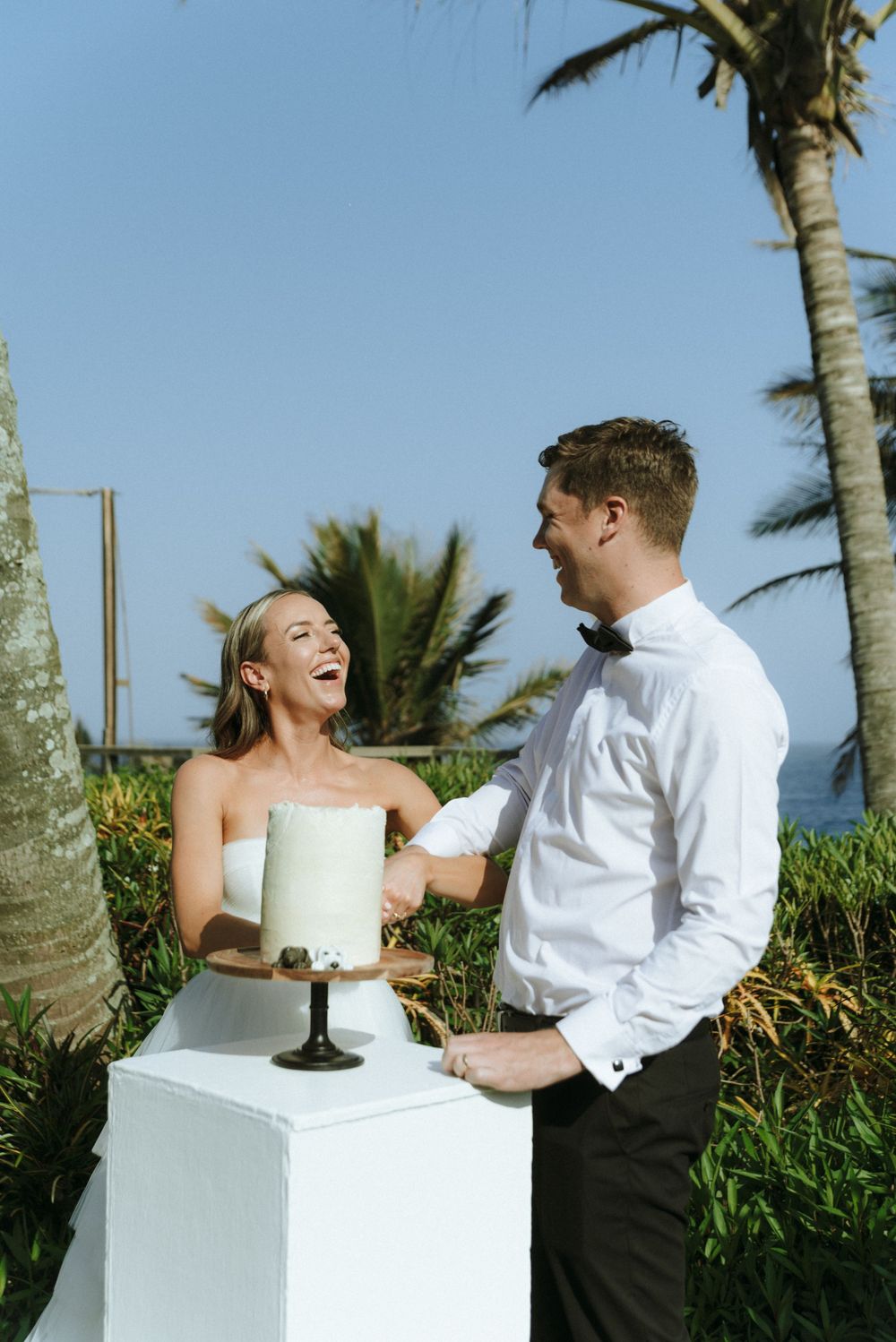 A couple shares a playful moment while cutting their wedding cake outdoors with palm trees in the background.