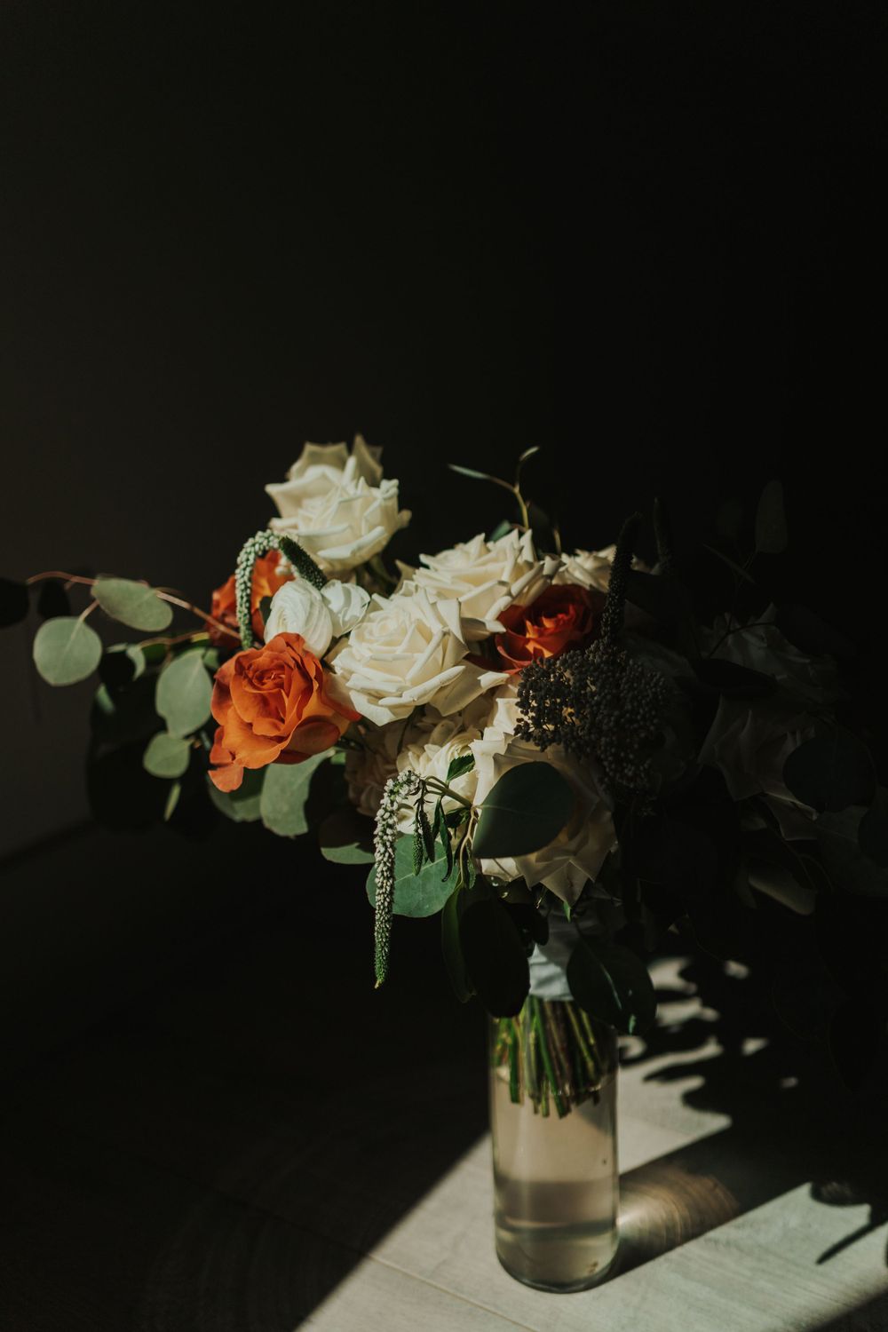 Dark and moody floral arrangement with white and orange roses in a clear vase casting dramatic shadows.