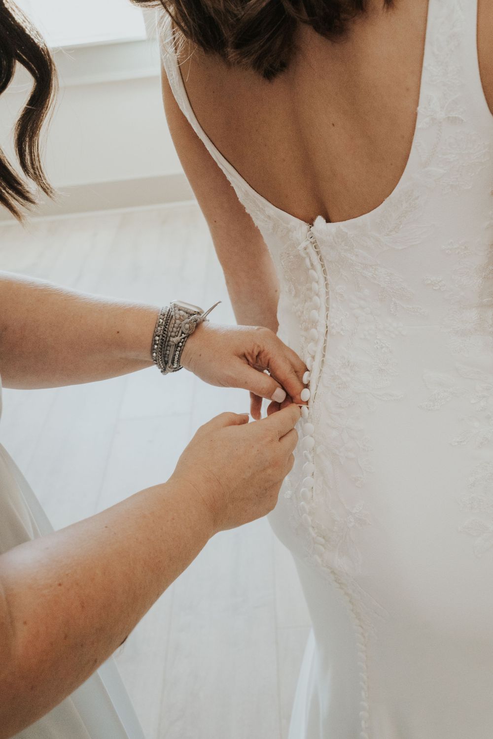 Close-up of hands buttoning white wedding dress from behind.