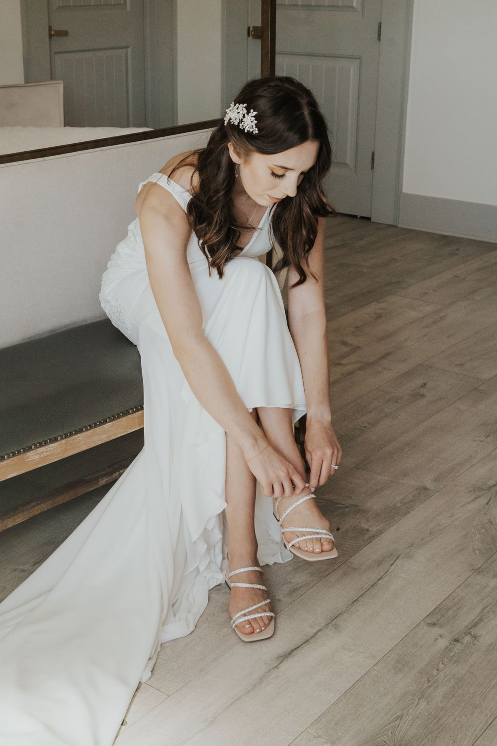 Bride in white dress adjusting strappy sandals while sitting on wooden floor.
