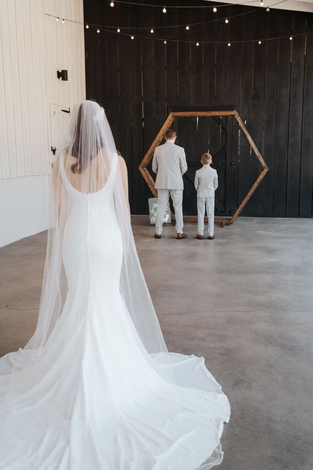 Bride in flowing veil watching two figures through hexagonal arch with string lights.
