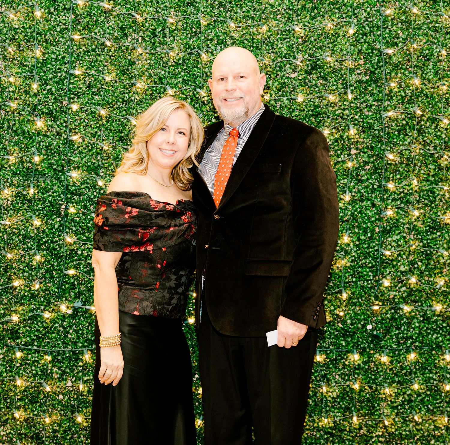A couple poses together against a green hedge wall with twinkling lights at a formal event.