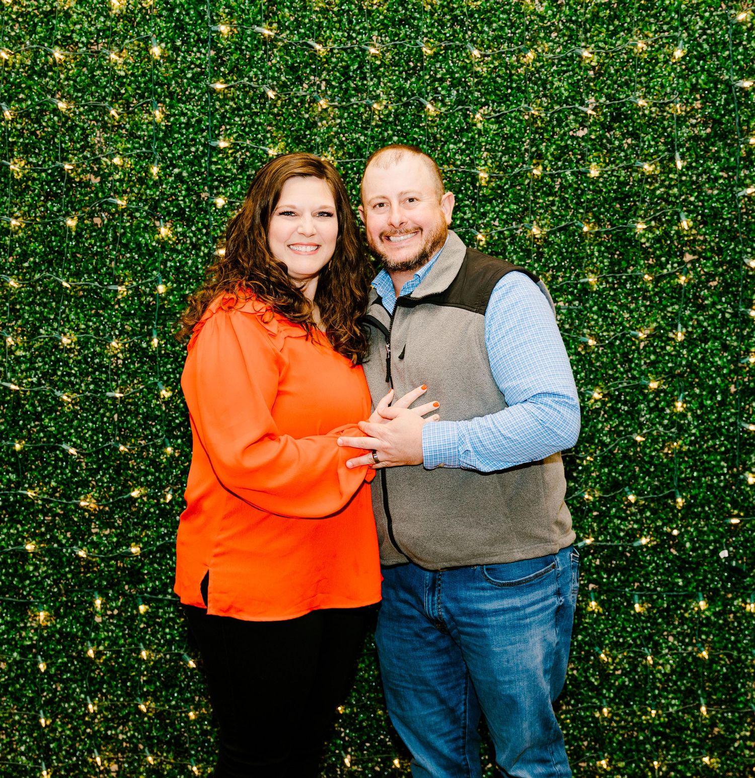 Couple poses together against a green hedge wall decorated with twinkling lights for a festive portrait.